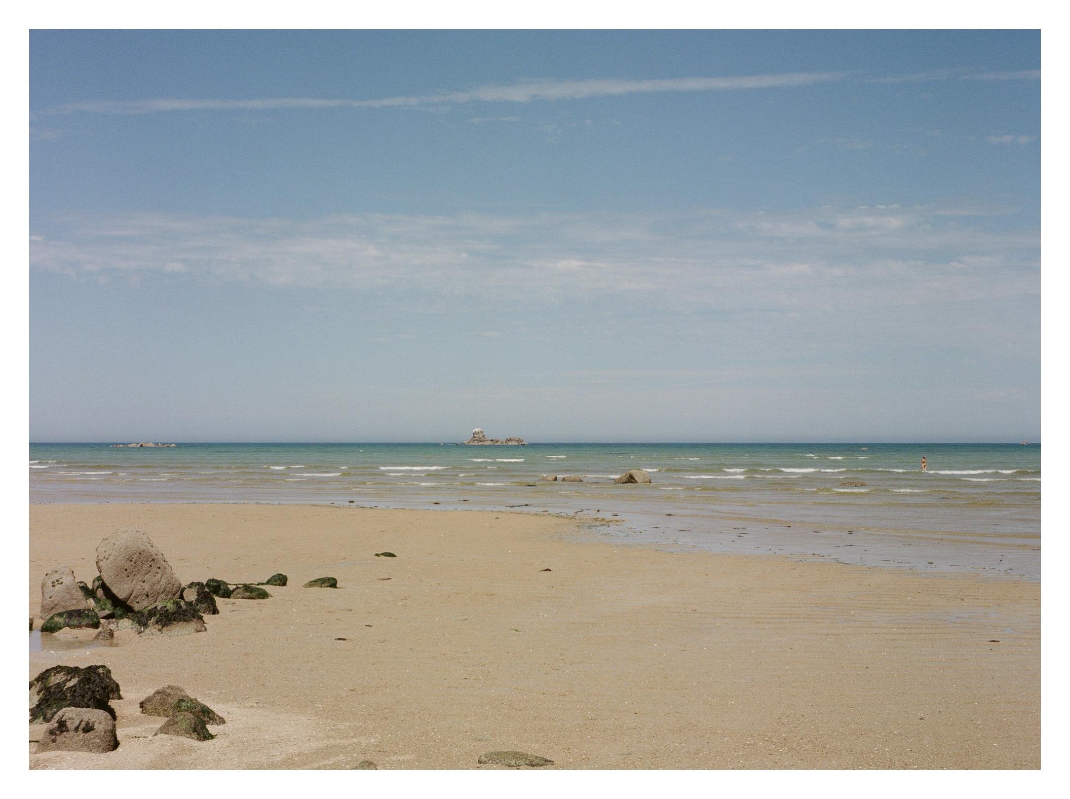 Wide sandy beach at low tide with scattered rocks in the foreground, shallow rippled water leading to a distant rocky outcrop near the horizon under a blue sky with thin clouds.