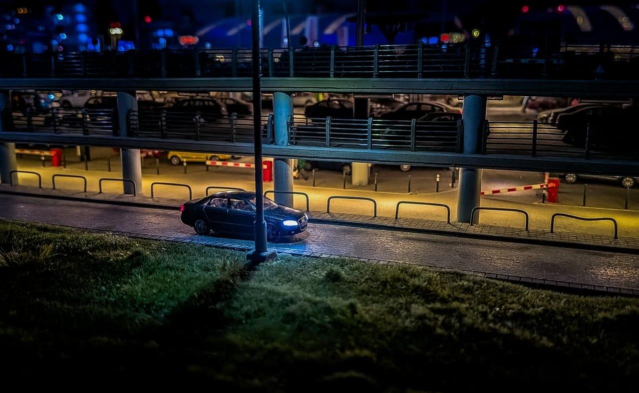 Black car parked along a curb at night beside a grassy slope and a streetlight pole, with a multi-level parking garage in the background lit by blue and yellow lights and lined with railings and barrier gates.