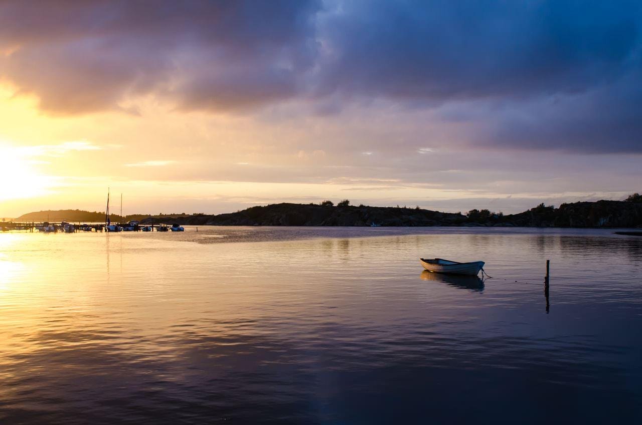 Small rowboat tethered on calm water near a wooden post, with distant anchored boats and a low shoreline under a partly cloudy sunset sky.