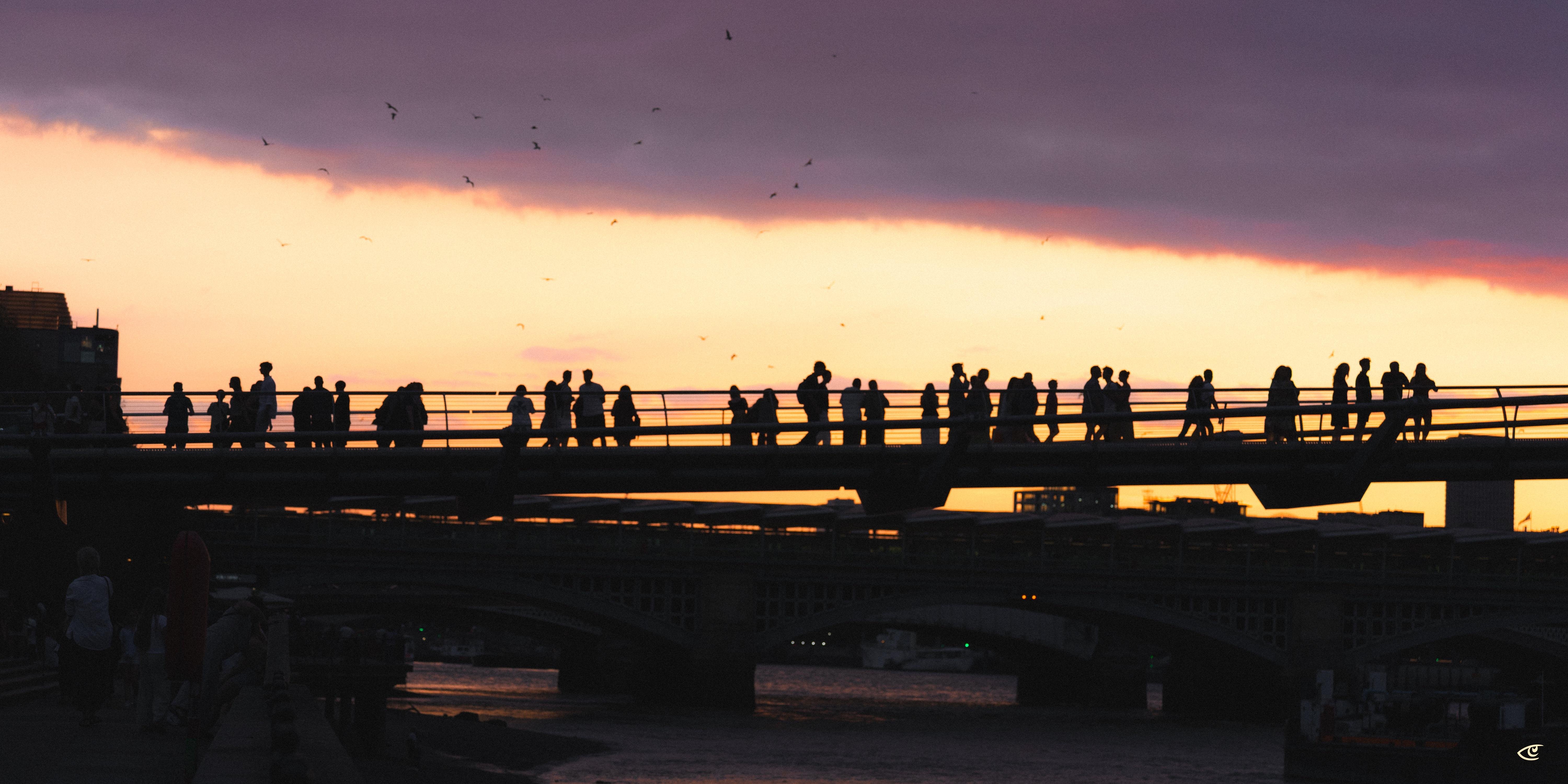 Silhouettes of people standing and walking along a bridge railing at sunset, with a dark cloud band overhead and an orange sky above a river and city buildings.