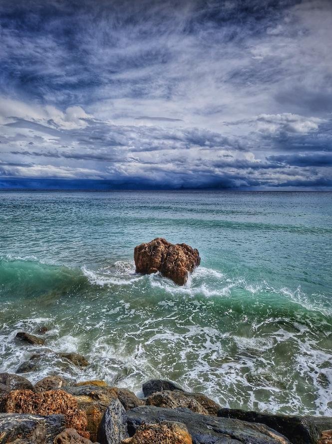 The image shows a seascape with a rocky coastline. There's a large rock in the foreground with waves crashing against it. The ocean water is a mix of turquoise and deep blue, and the sky is filled with dramatic, textured clouds, suggesting an overcast day or an approaching storm.