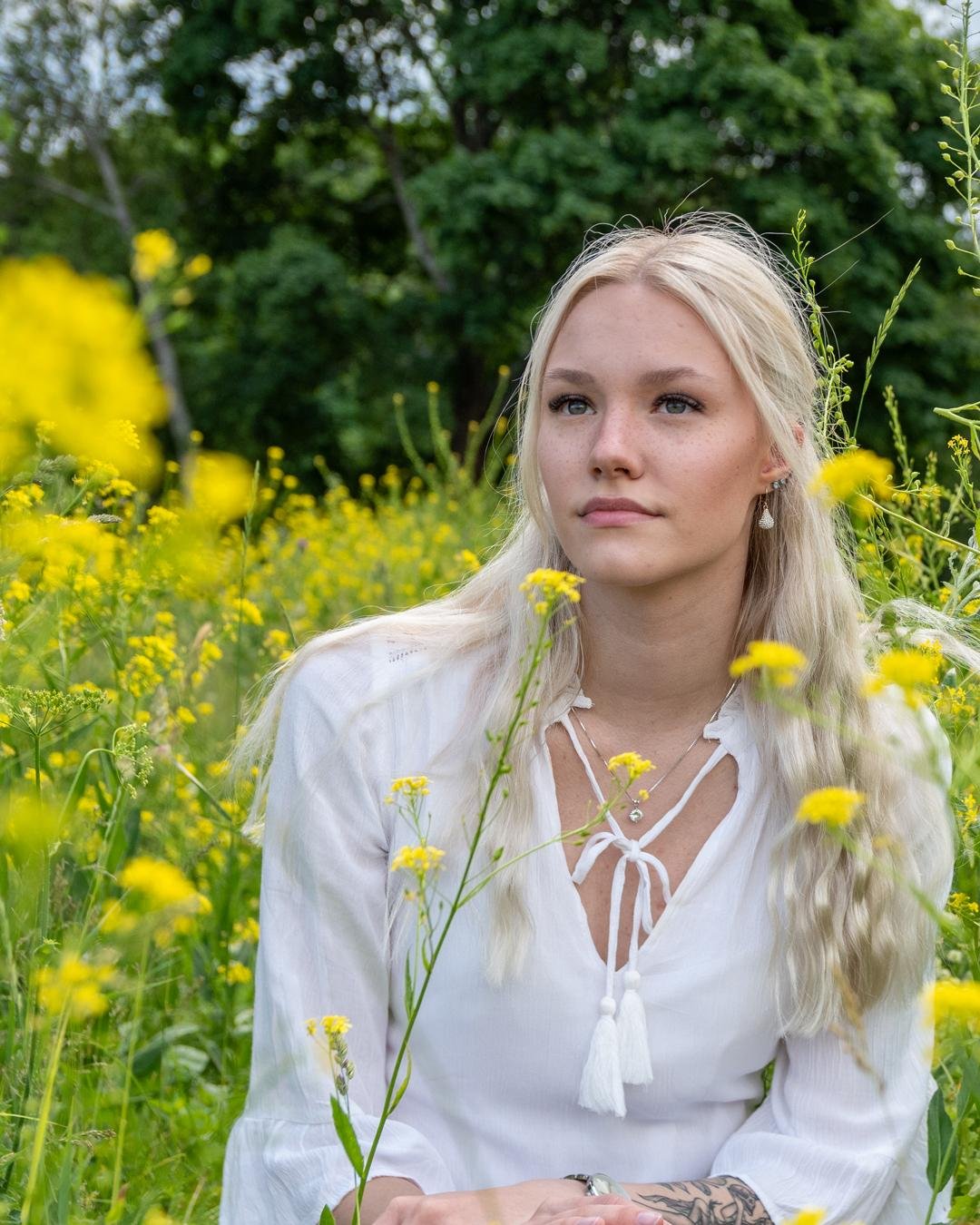 The image shows a person with long blonde hair sitting in a field of yellow flowers. They are wearing a white top and there are green trees in the background.