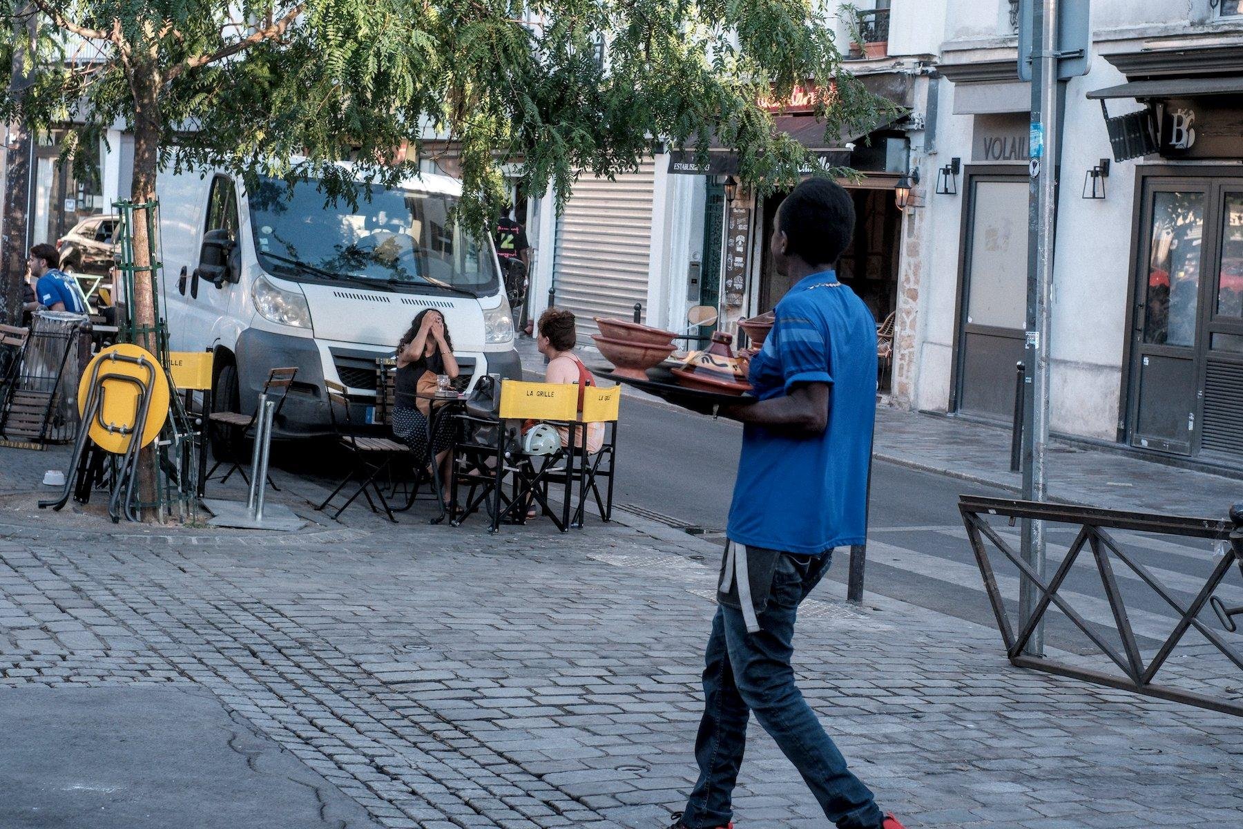 Photo horizontale. Sure une place pavée parisienne, un homme en haut bleu et jean, transporte un plateau plein de plats exotiques vers la terrasse du restaurant sur la place