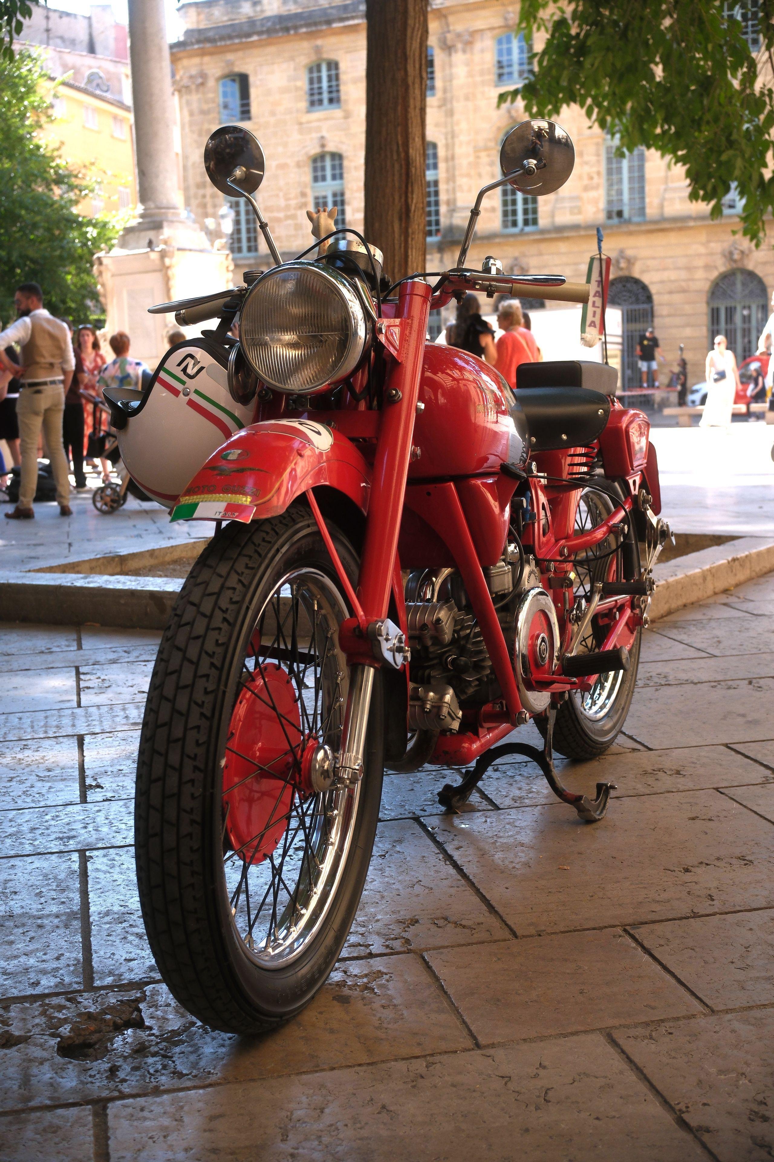 Photo format portrait prise accroupie vu l’angle, montrant une moto rouge de collection de 3/4 face, au look vintage, avec un casque blanc style ancien aussi pendu au guidon droit.