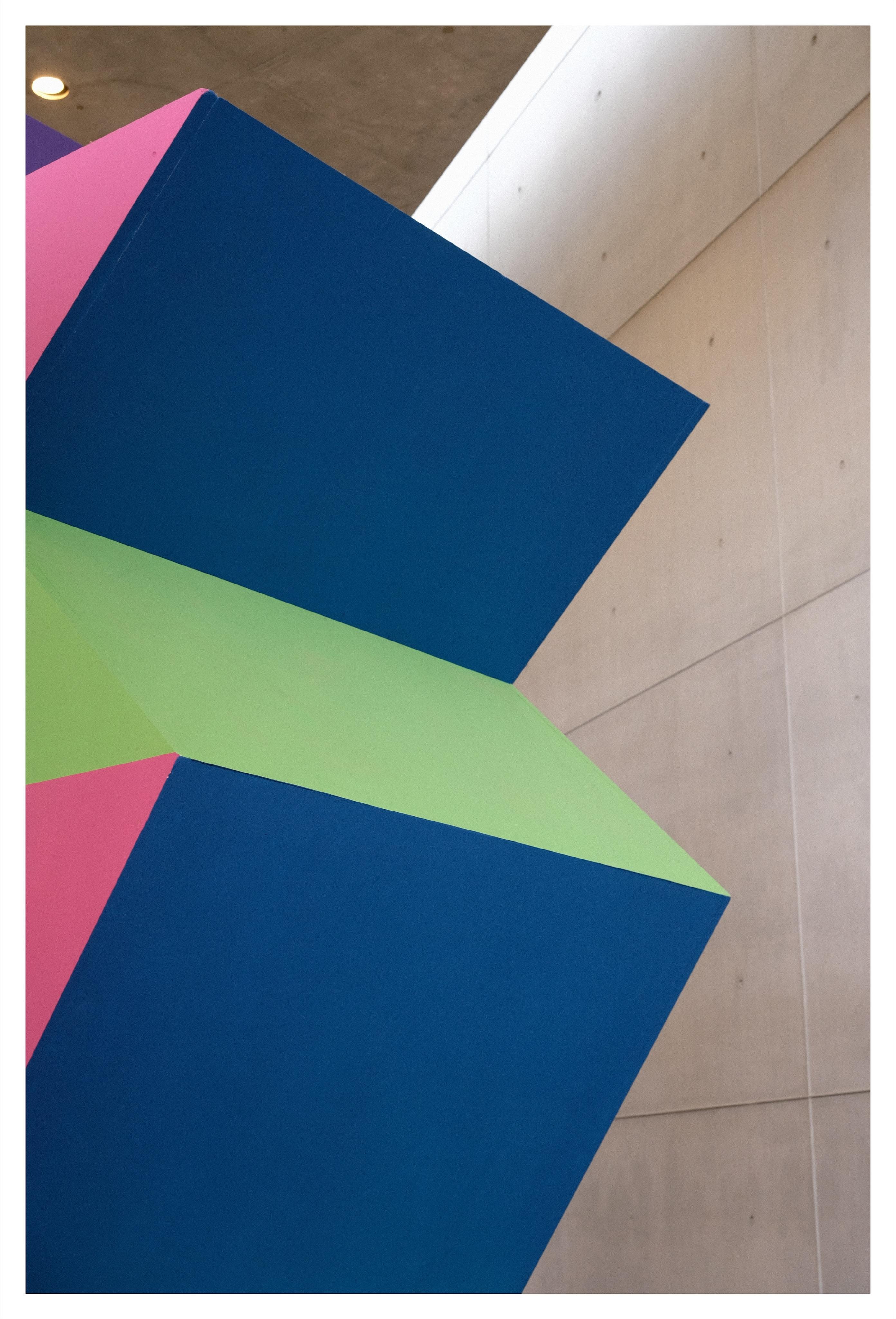 A wooden sculpture made out of flat surfaces and sharp corners, coloured in bright green, blue and pink, in front of a flat concrete wall.