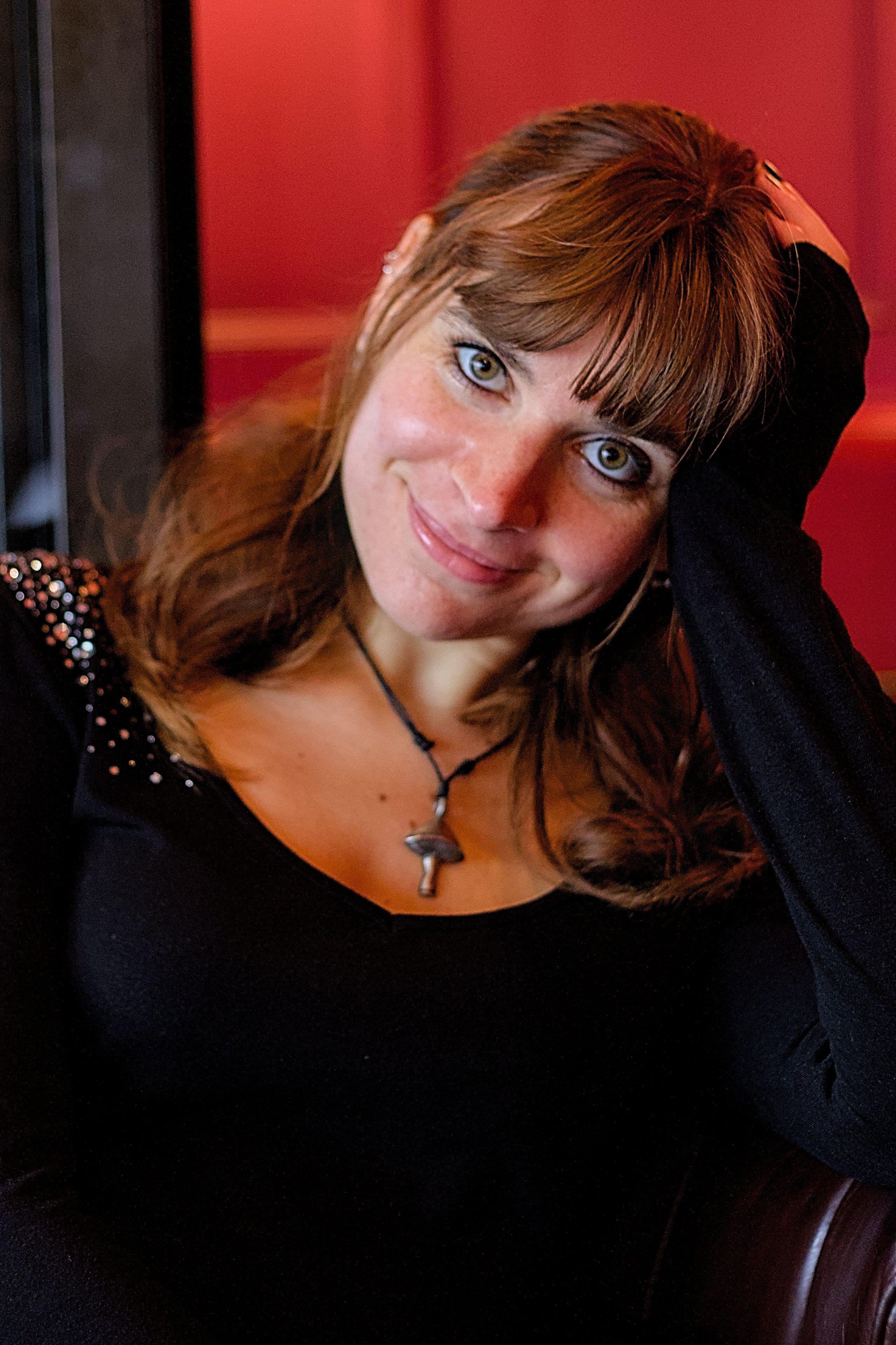 Photo format portrait couleur, montrant une jeune femme brune au cheveux long, habillée en noir, posant sa tête dans sa main gauche, le bras accoudé sur une chaise. Le mur derrière est rouge vif, contrastant avec sa tenue noire. Elle regarde la caméra avec ses yeux vert et souri. Elle porte aussi un pendentif en forme de champignon