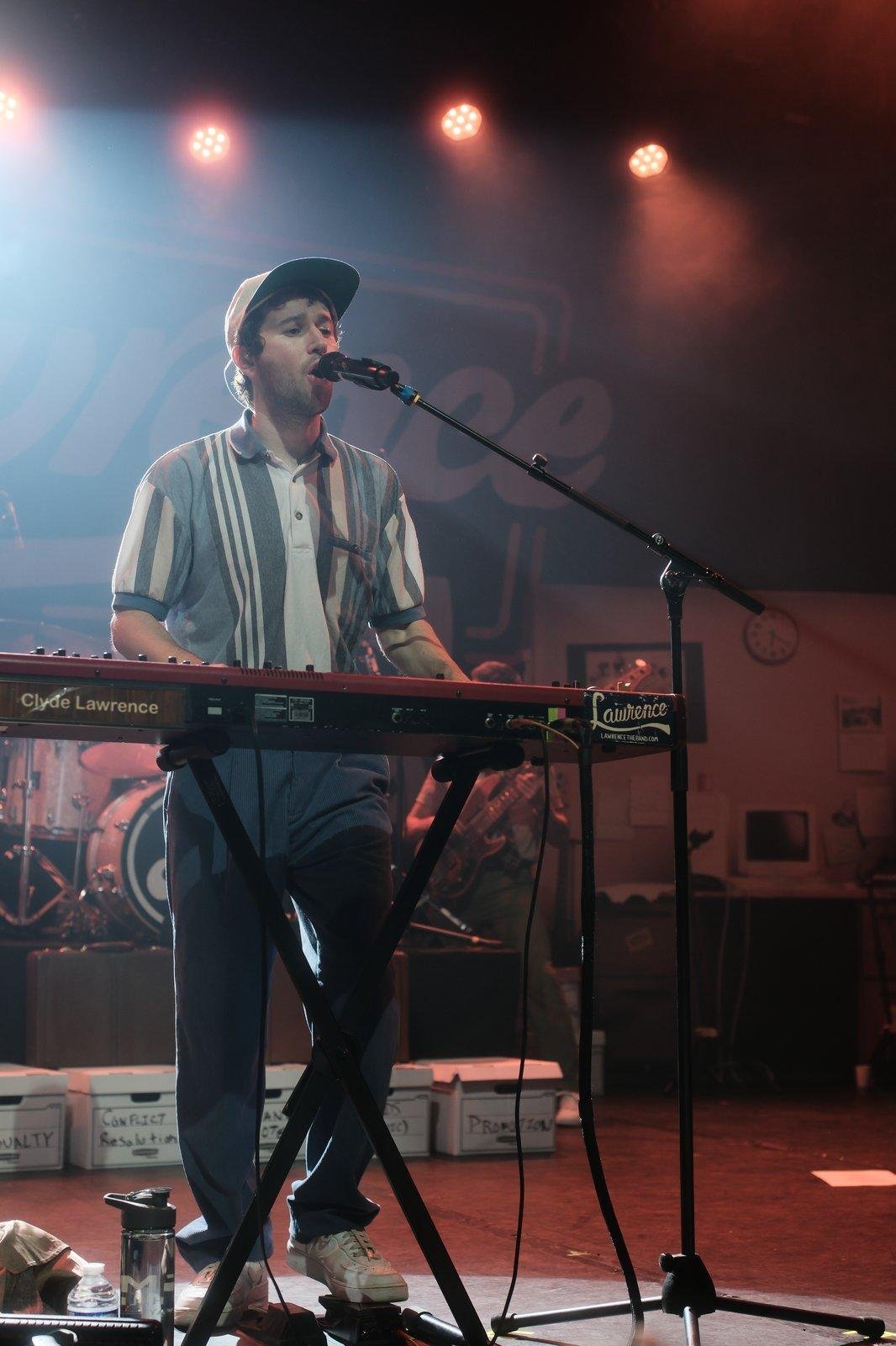 Photo verticale. un jeune homme derrière un clavier de piano électronique et un micro. Portant une casquette, une chemise et un