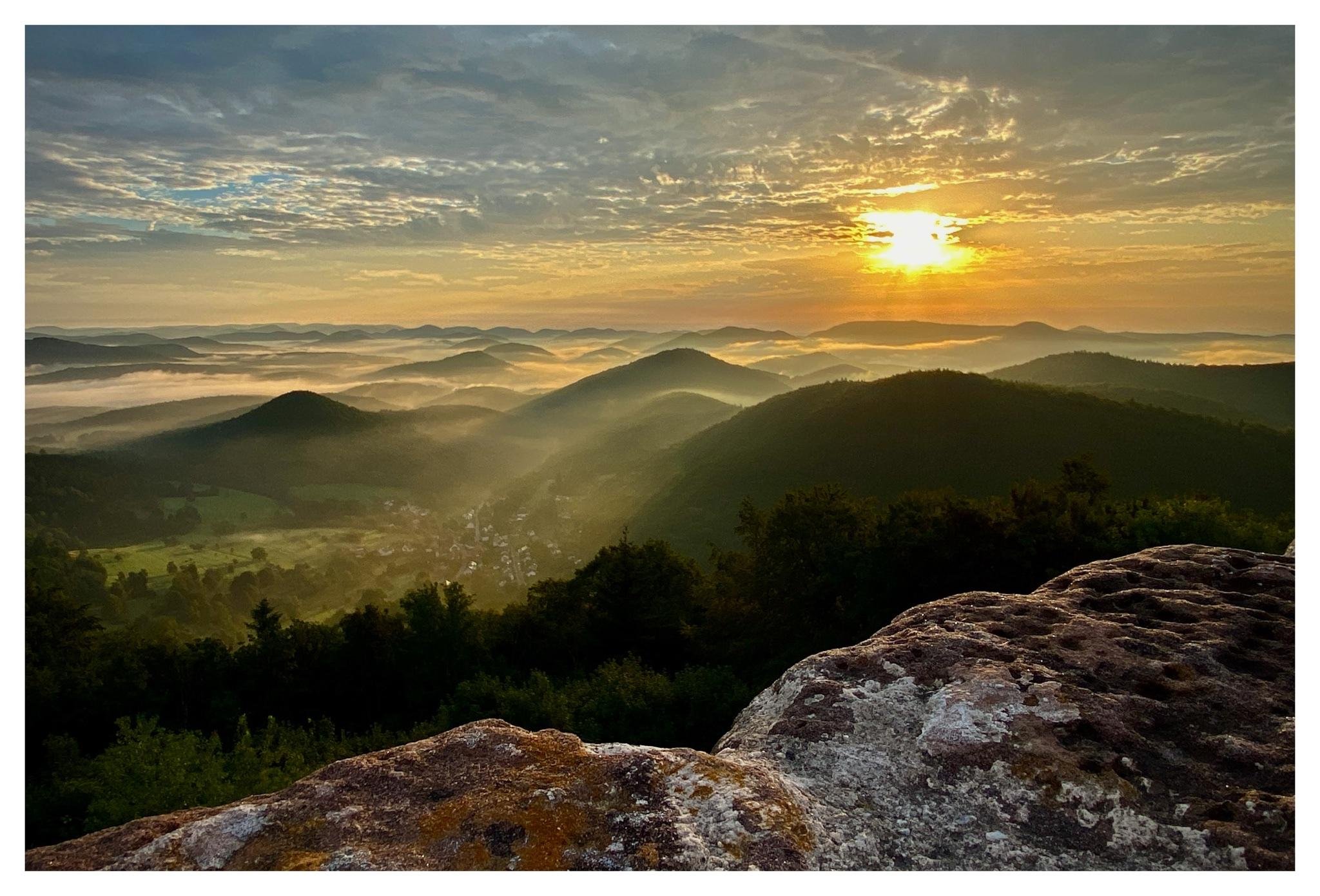 Sunrise over layered, forested hills with mist filling the valleys; a rocky ledge in the foreground and a partly cloudy sky with the sun near the horizon.