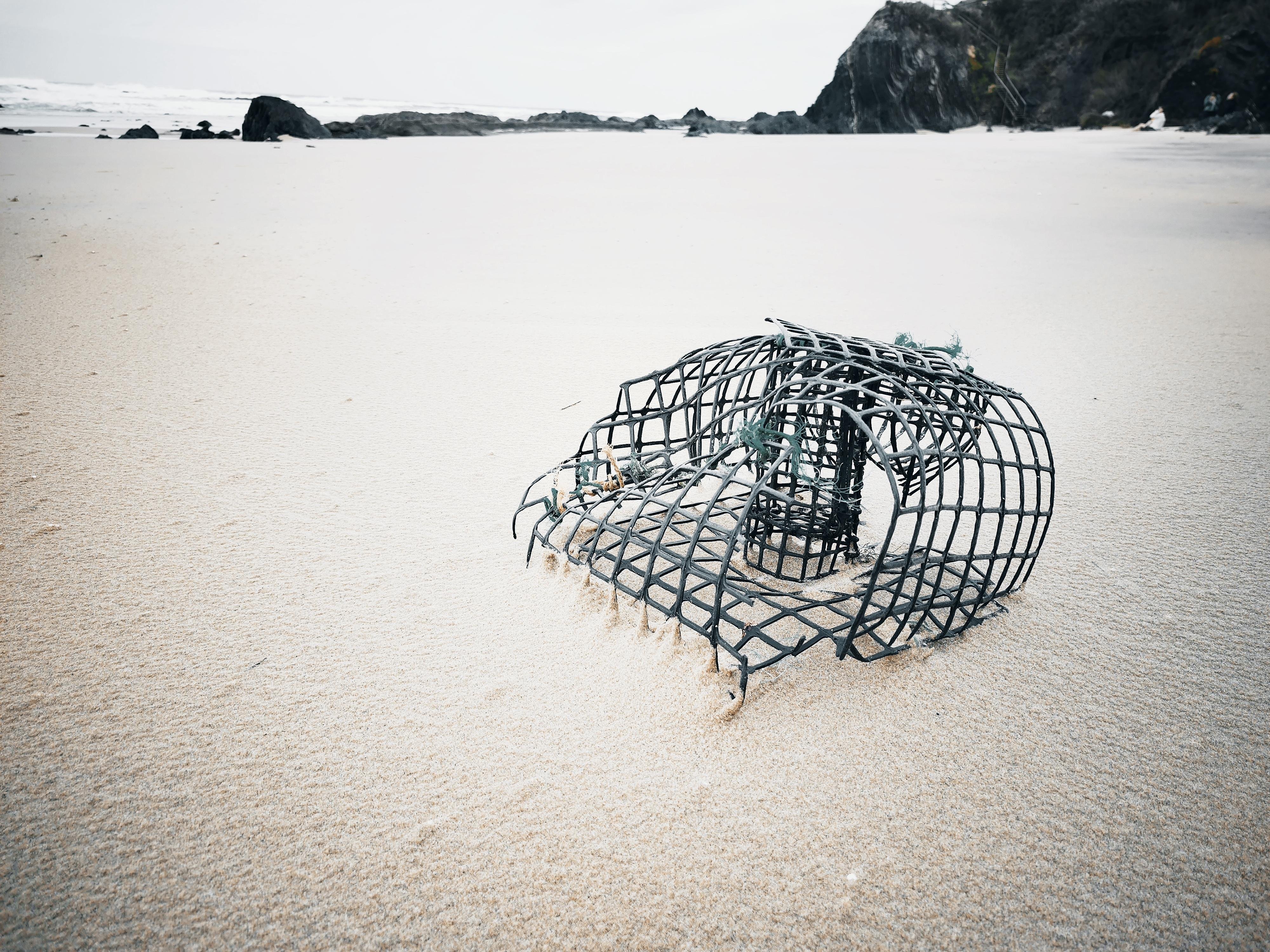 This image depicts an empty lobster or crab cage lying on a sandy beach. The cage appears slightly bent or damaged, and the scene has a calm, overcast atmosphere with a distant view of rocky outcroppings and the ocean.