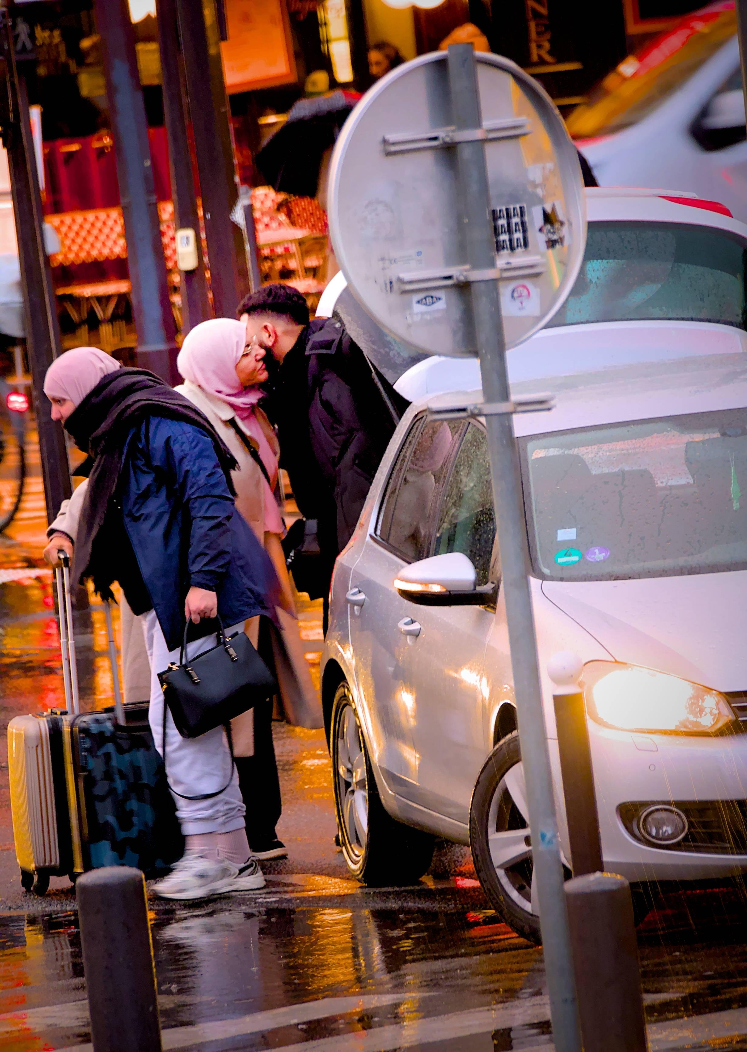 La photographie représente une scène de rue avec trois personnes à côté d'une voiture dont le coffre est ouvert. Deux femmes portant un foulard portent des bagages, l'une d'elle fait la bise à un homme. C'est un jour de pluie, comme le montrent la rue mouillée et les reflets. Un panneau de signalisation se trouve également au premier plan.