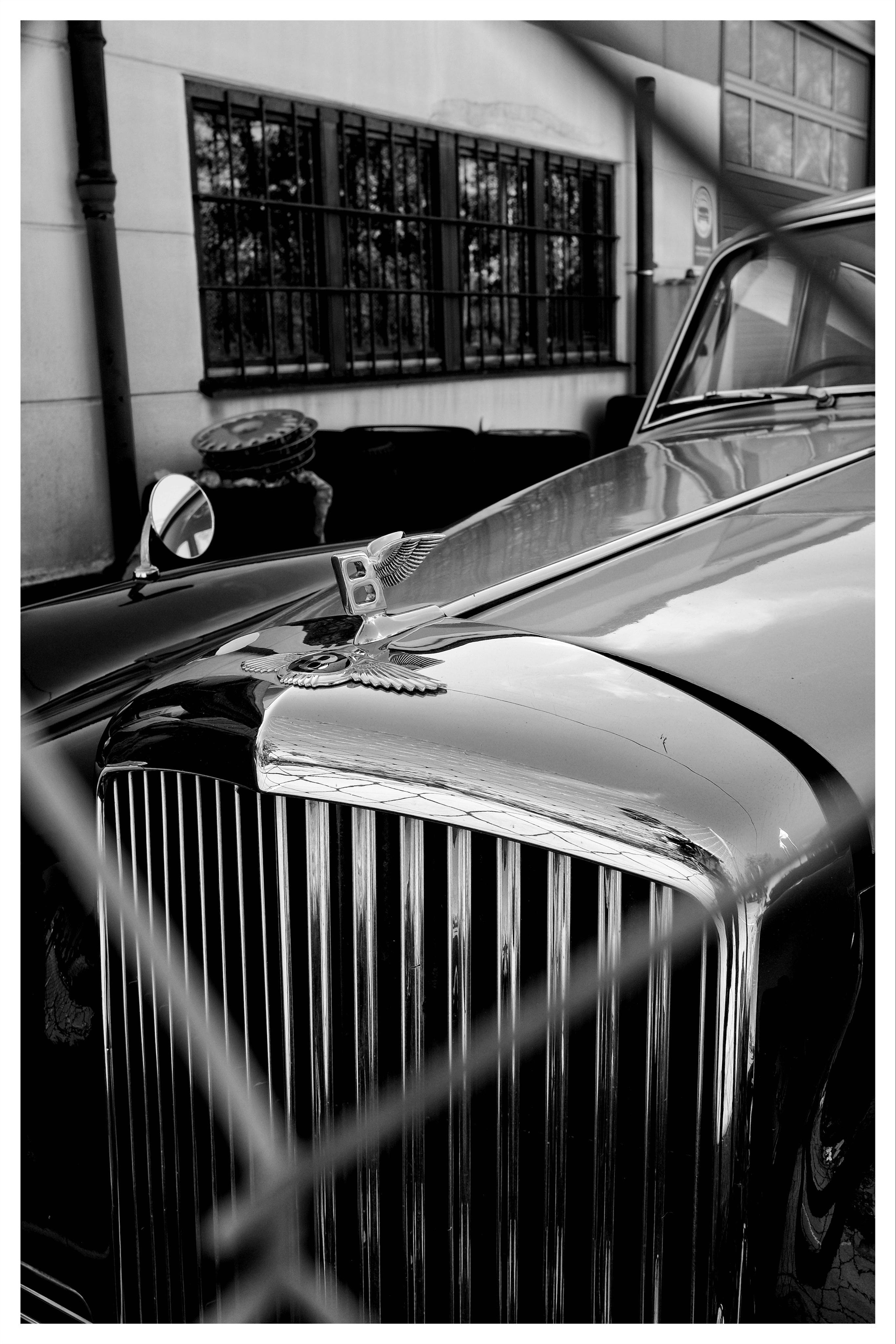 Black-and-white close-up of a classic car’s front end, showing a winged hood ornament, chrome grille, and side mirror, with a building’s barred windows in the background and blurred fence lines crossing the foreground.