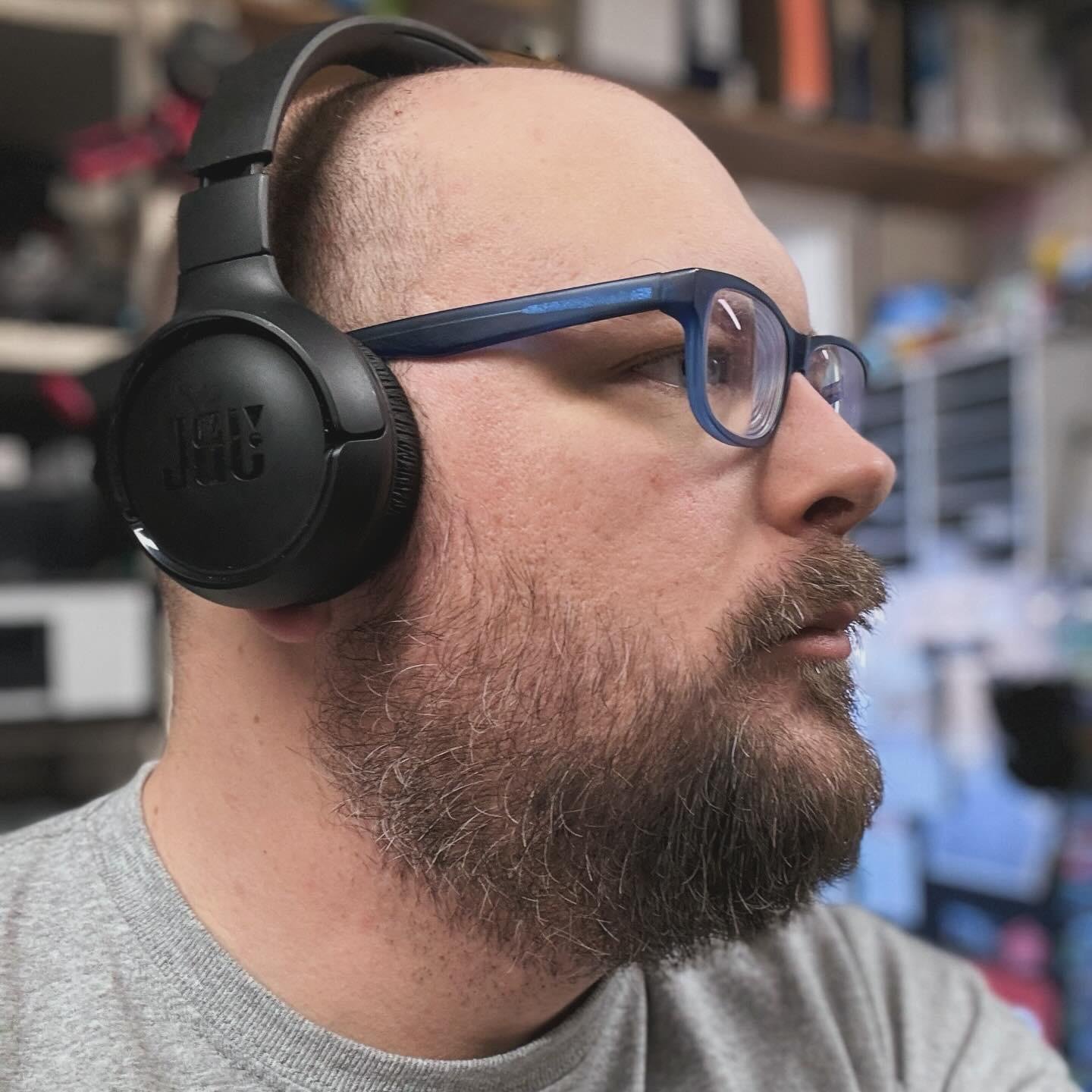 The image shows a person wearing headphones and glasses. The person has a beard and is looking to the side. The background appears to be an indoor setting, possibly an office or a room with various items on shelves.