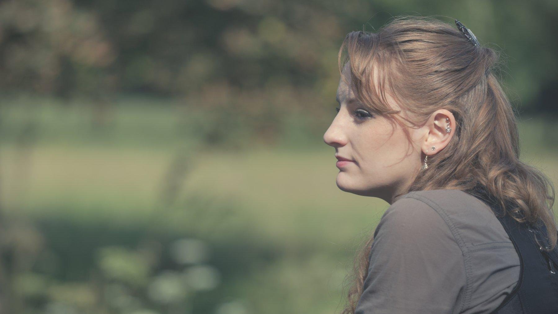 Photo horizontale. Portrait d’une jeune femme rousse portant un chemisier marron et un corset noir, de profile, et semble accoudée. Elle regarde droit devant elle. Le fond est flou fait de verdure. La lumière est un peu glowy