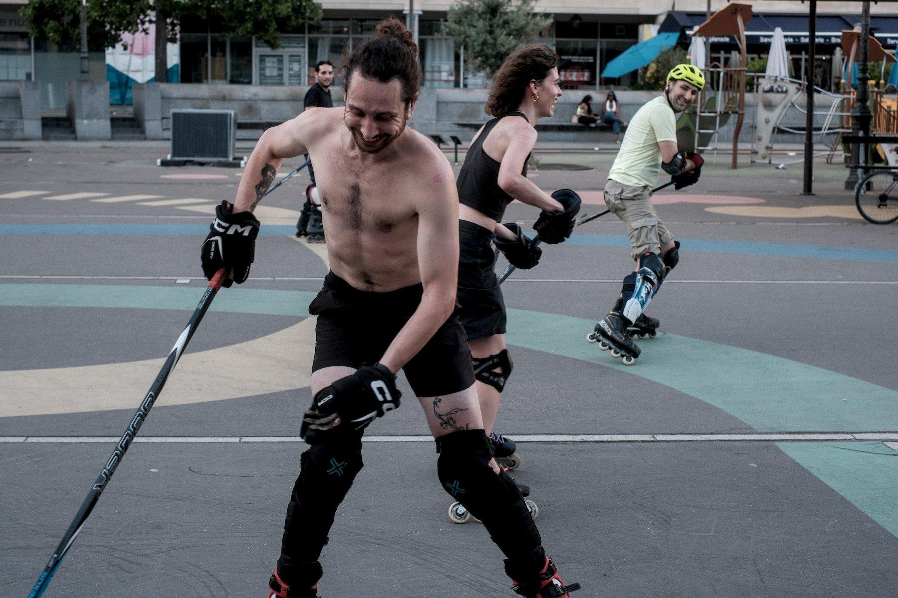 Photo horizontale. Au premier plan, un homme torse nu, en short noir, protège tibia, gants de hockey, crosse de hockey dans la main droite et on voit le haut de ses roller. Il rit en regardant vers le bas sûrement en train de récupérer le palet. Derrière, une jeune femme en débardeur noir short noir, gants de hockey, crosse à deux main, genouillères et roller, riant aussi en train de partir se repositionner. Encore sur le plan d’après, un homme, casque de vélo, t-shirt vert pâle, short cargo beige, genouillères et protège tibia, roller, riant aussi et partant se repositionner aussi