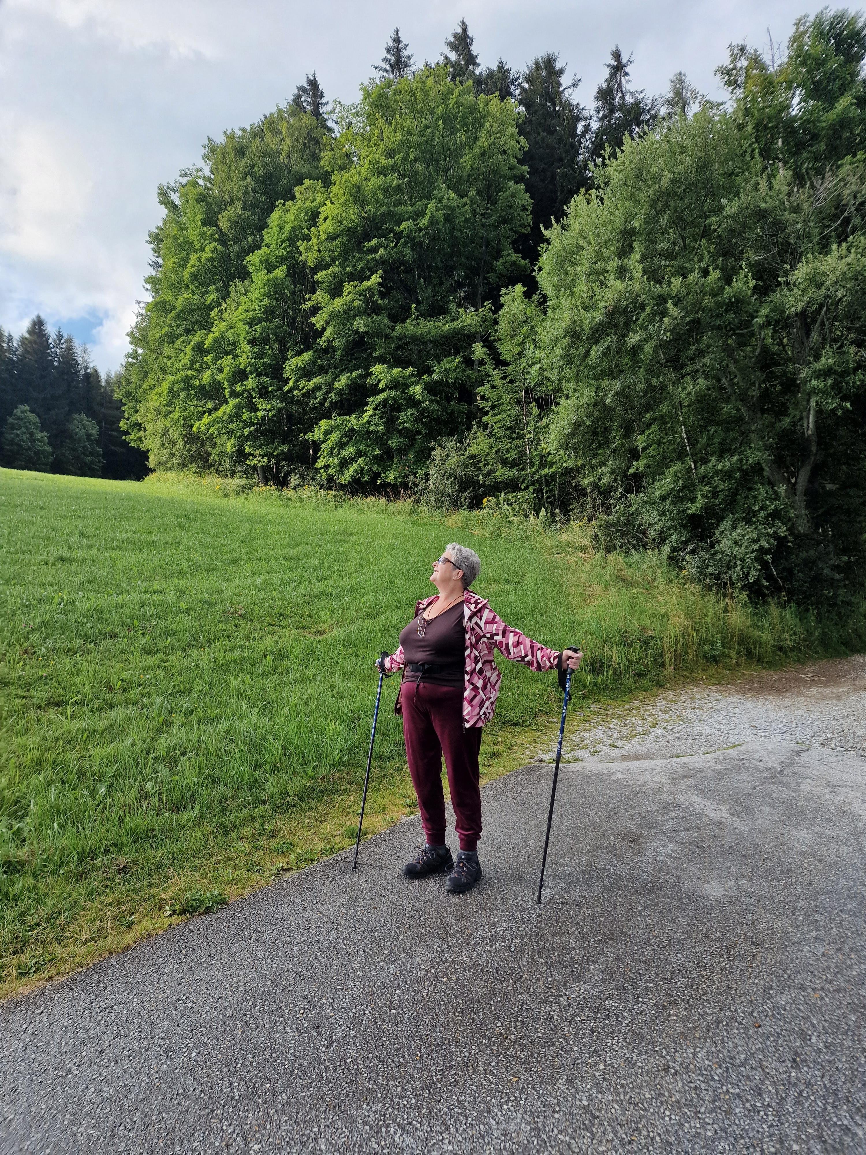 Cynni’s mum stands on a quiet mountain path in Brandlucken, Steiermark, Austria, during a peaceful morning walk after the long 1100-kilometer car journey the day before. She faces the soft light breaking through the clouds, arms stretched wide with her hiking poles in hand, as if welcoming the warmth of the sun. Her maroon walking clothes and patterned jacket contrast beautifully with the vivid green meadow beside her and the dense forest rising behind her. The scene feels calm and joyful—nature still damp from the morning, air fresh, and sunlight gently touching everything. Her expression is relaxed and content, capturing a rare, tender moment of pure happiness. Surrounded by Austrian hills, forests, and the quiet of early day, this photo radiates warmth, love, and the simple joy of being together in a beautiful place.