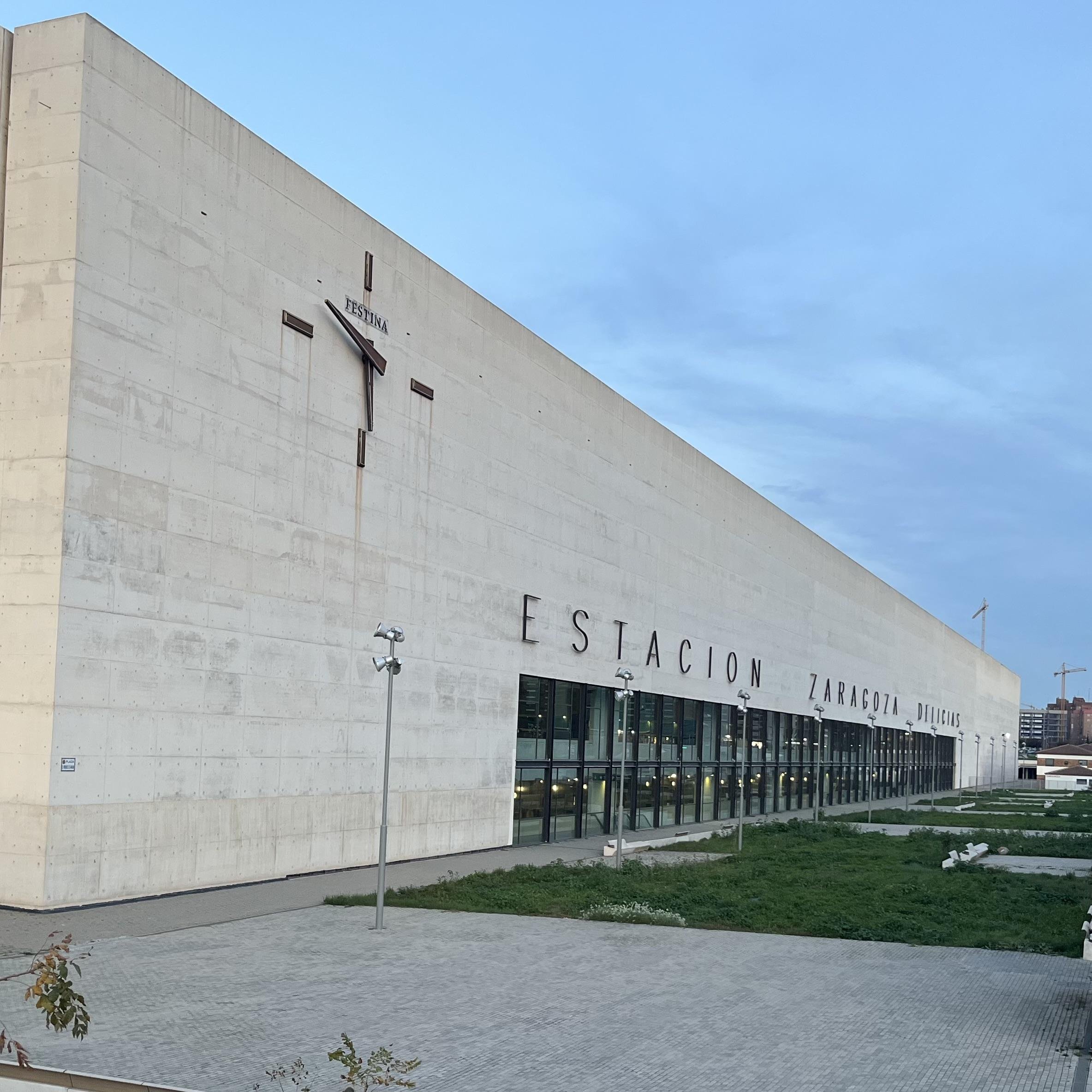 The image shows the exterior of the Zaragoza Delicias Station in Zaragoza, Spain. The building is modern with a large inscription "ESTACION ZARAGOZA DELICIAS" on its facade. The architecture is minimalist, made of concrete, and there are glass windows and some greenery in front of the structure.