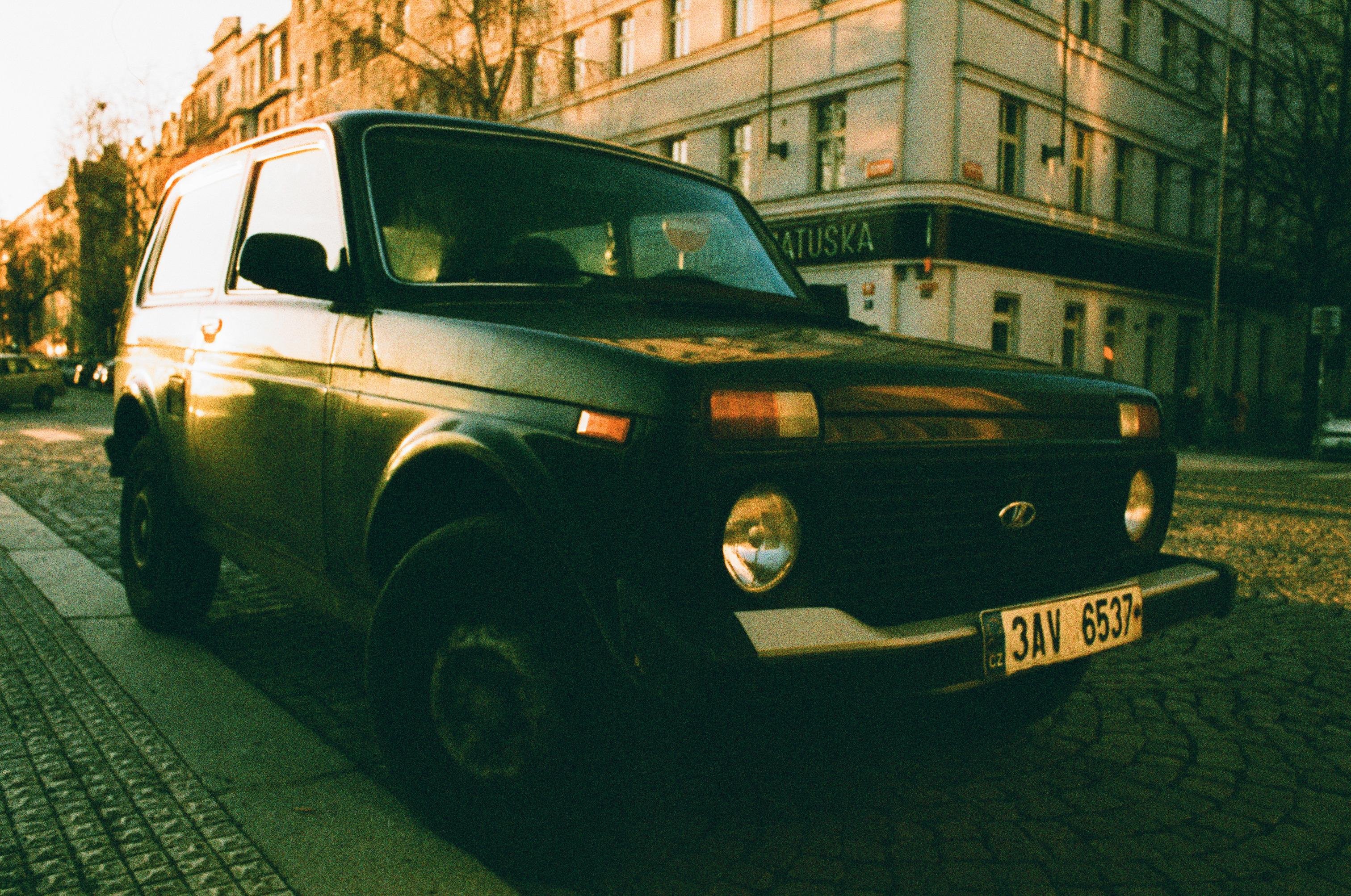 A yellow tone photo of a black 70's era soviet offroad car parked on a street with european style interwar buildings.