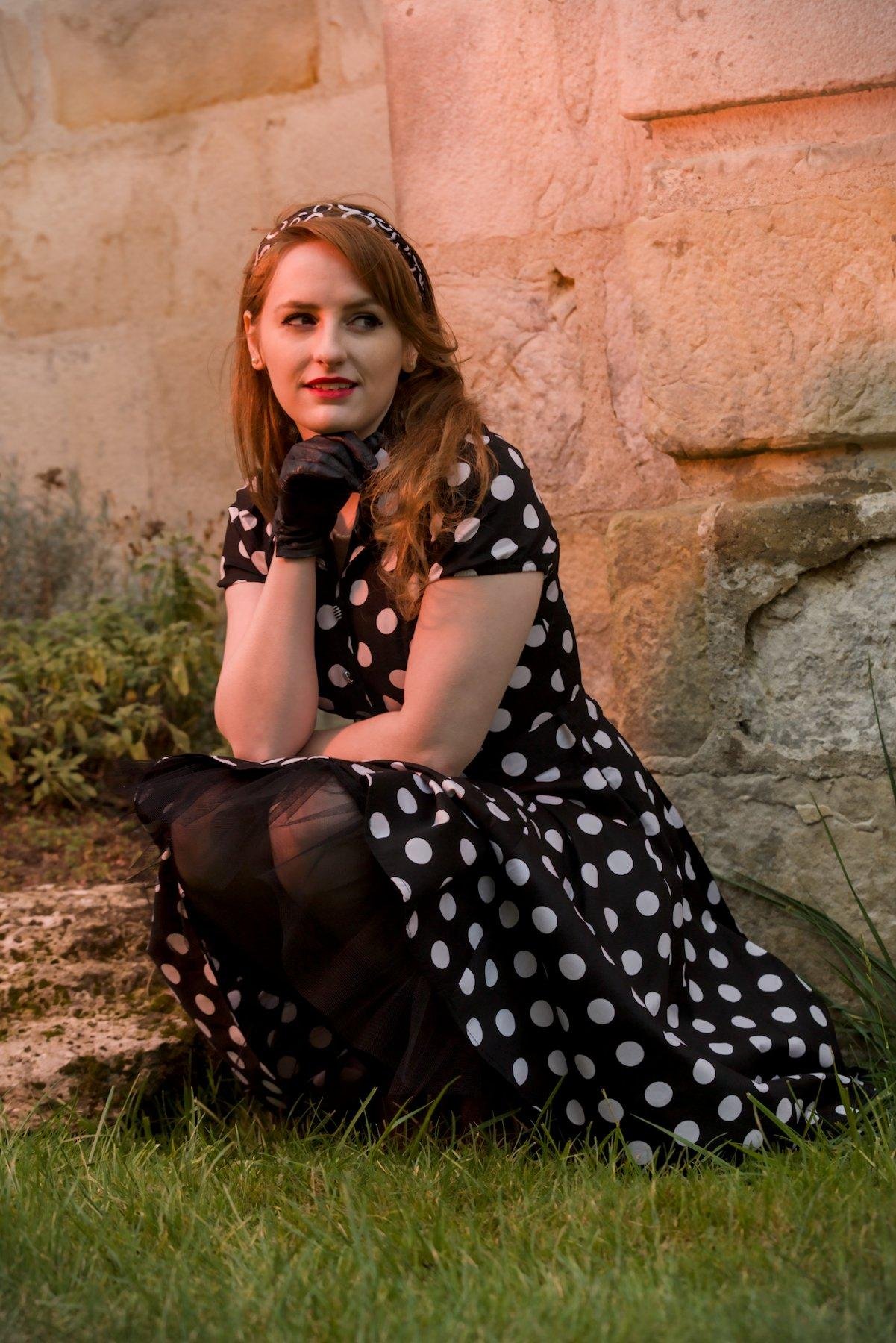 Photo verticale. Une jeune femme aux cheveux long roux, posant devant un mur de vieilles pierres, portant une robe noire à pois blanc, des collants noirs, des chaussures noires avec un petit nœud, des gants  en cuir noir et un serre tête noir avec des motifs blancs. Elle est accroupie et pose sa tête sur le dos de sa main droite. Son bras gauche est posé sur ses genoux. Elle regarde à sa gauche en souriant