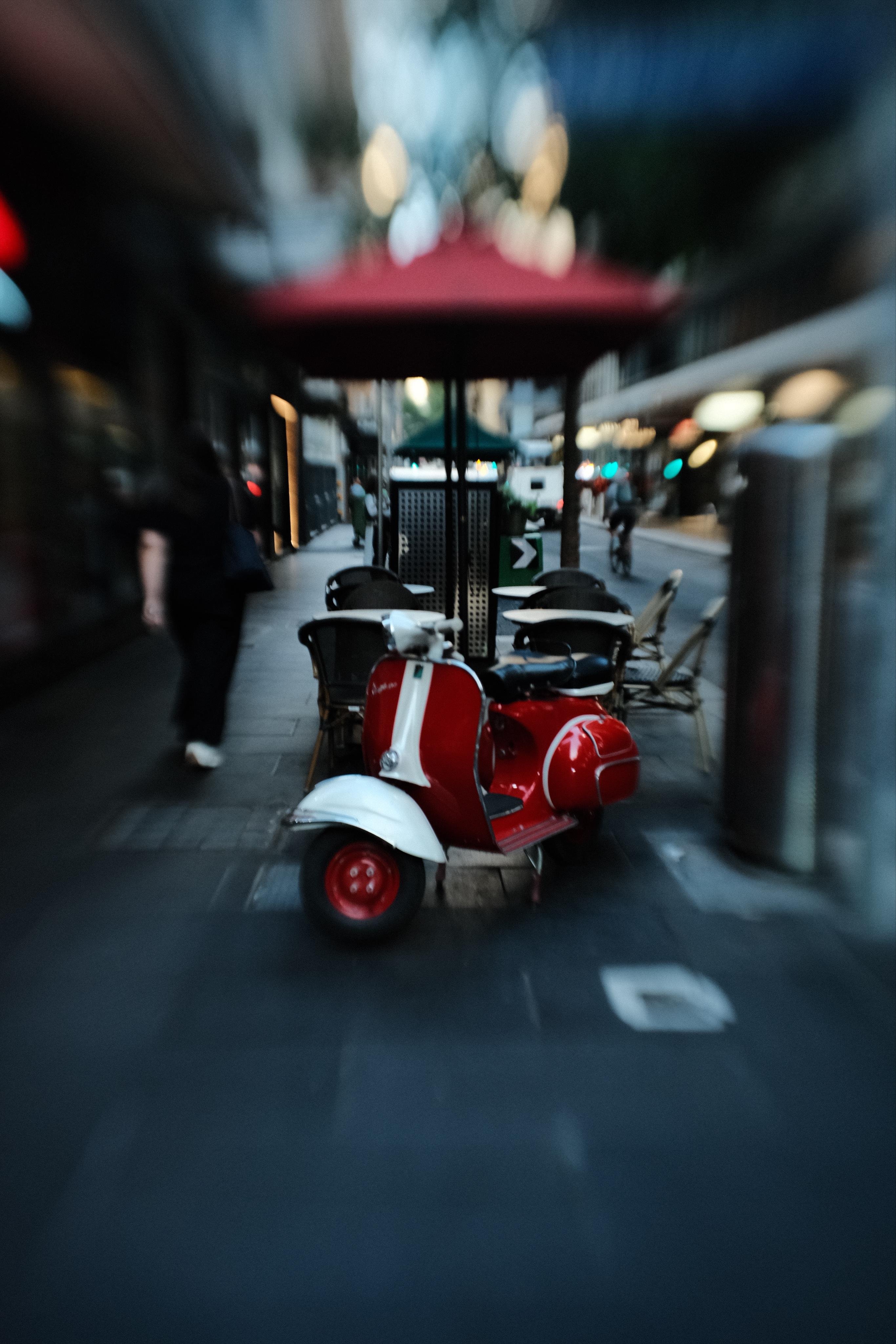 Red-and-white vintage scooter parked beside outdoor café tables under a red umbrella on a city sidewalk, with pedestrians and storefront lights blurred in the background.