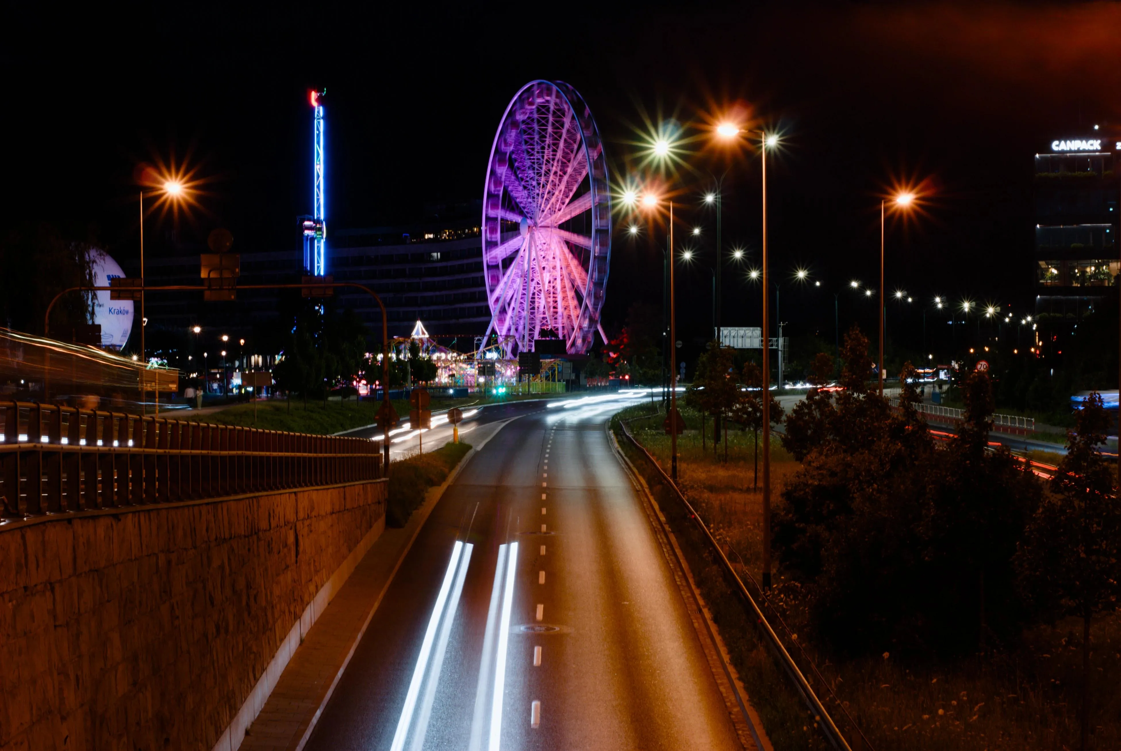 A photo of a ferris wheel at night with the light rays from the cars passing by on the road