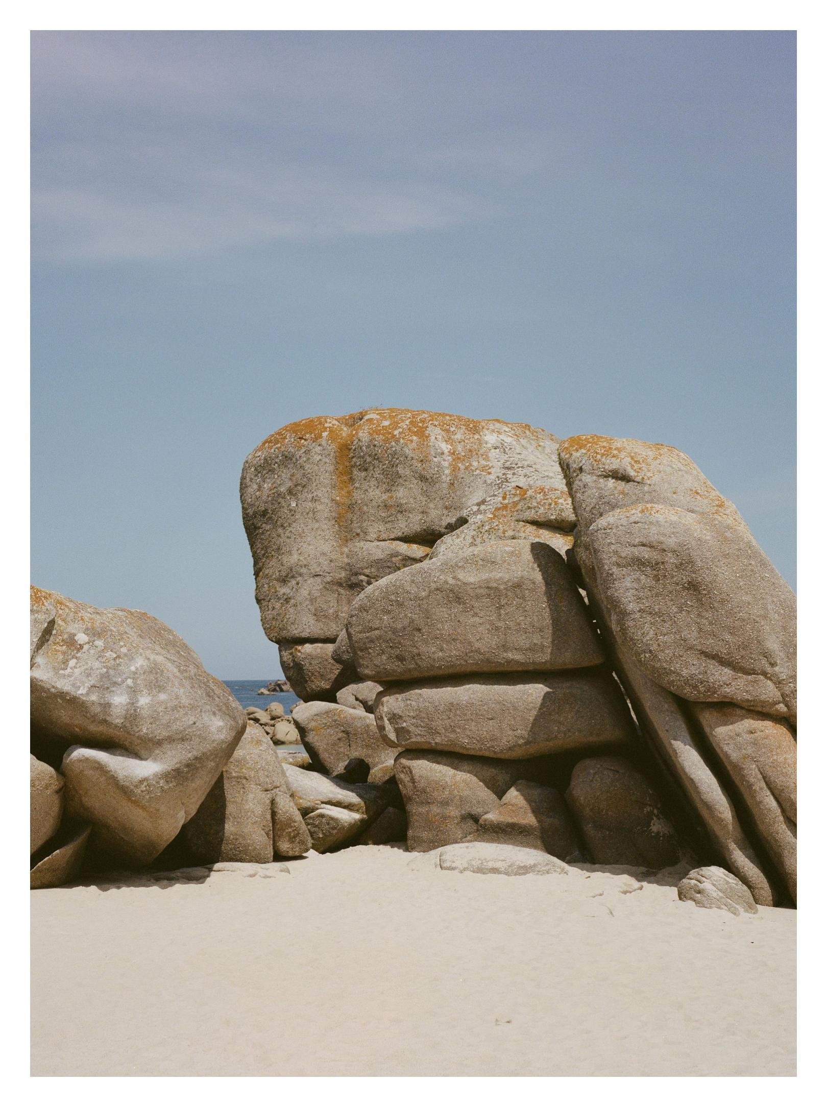 Large weathered granite boulders stacked on a sandy beach, with a strip of ocean visible between rocks and a clear blue sky above.