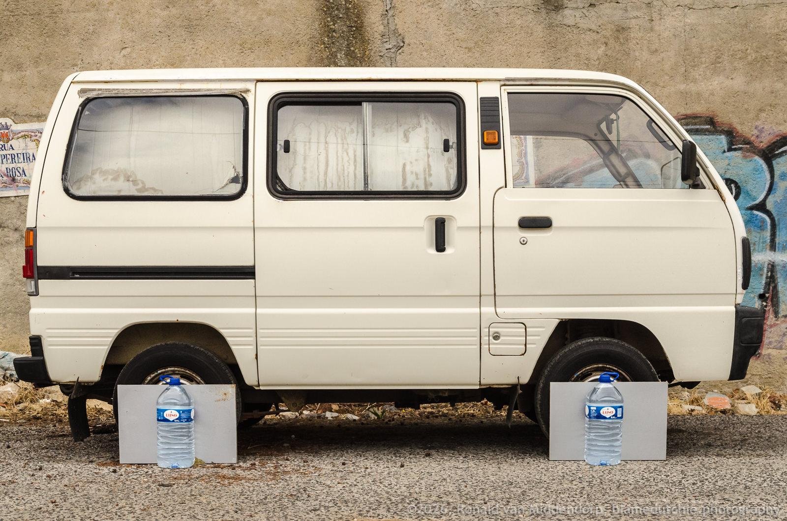 Side view of a white van parked on a street in front of a graffiti-covered concrete wall, with the front and rear wheels partially hidden by grey painted planks and large clear water bottles.