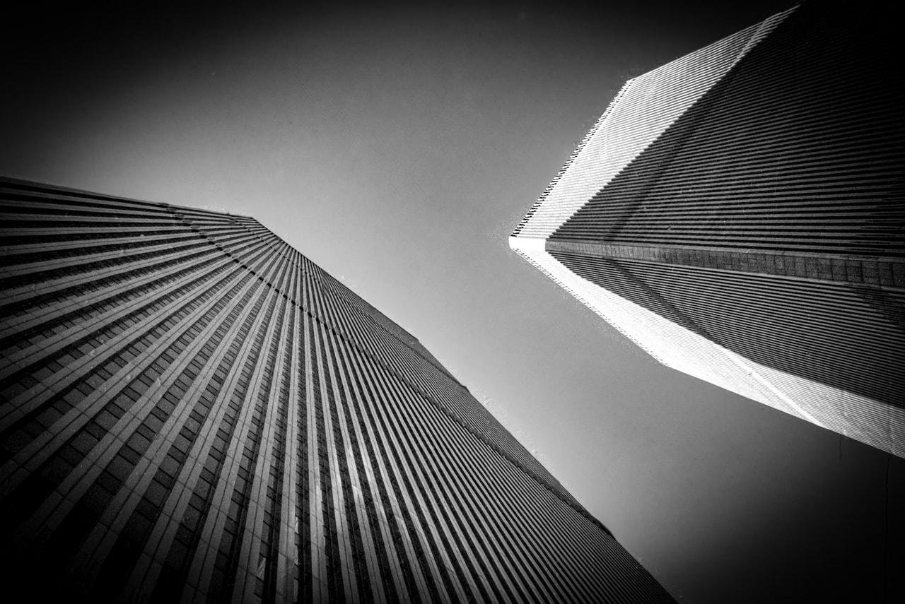 Black and white photograph of two skyscrapers viewed from a low angle. The buildings are prominent, featuring vertical lines and converging towards the top with a clear sky backdrop. The left building is closer, displaying more defined windows, while the right building shows strong diagonal shadows due to lighting. The composition emphasizes geometric shapes and contrast.
