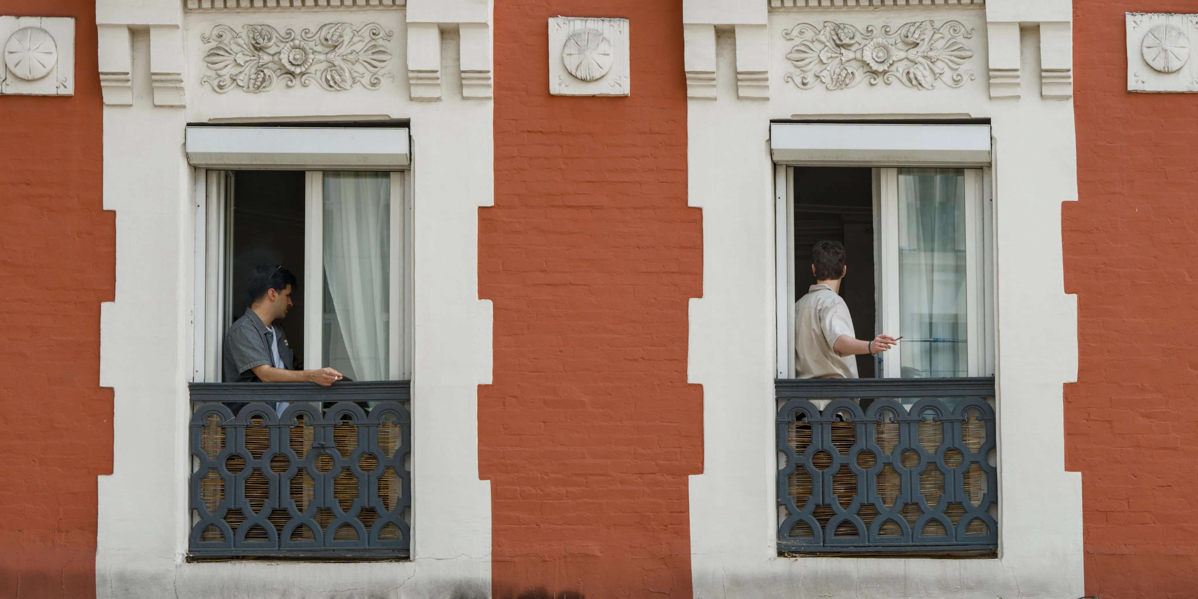 Photograph of two windows in a brick building. The building is painted orange-red. The decoration around the windows is white. The windows each have iron railings, doubled with a bamboo mat. The windows are half open on the same side. In each one of the two windows there is a man turning his head to the inside of the room, looking away from the camera. The men are both smoking cigarettes, extending their left arm holding the cigarette outside their respective windows over the railings.