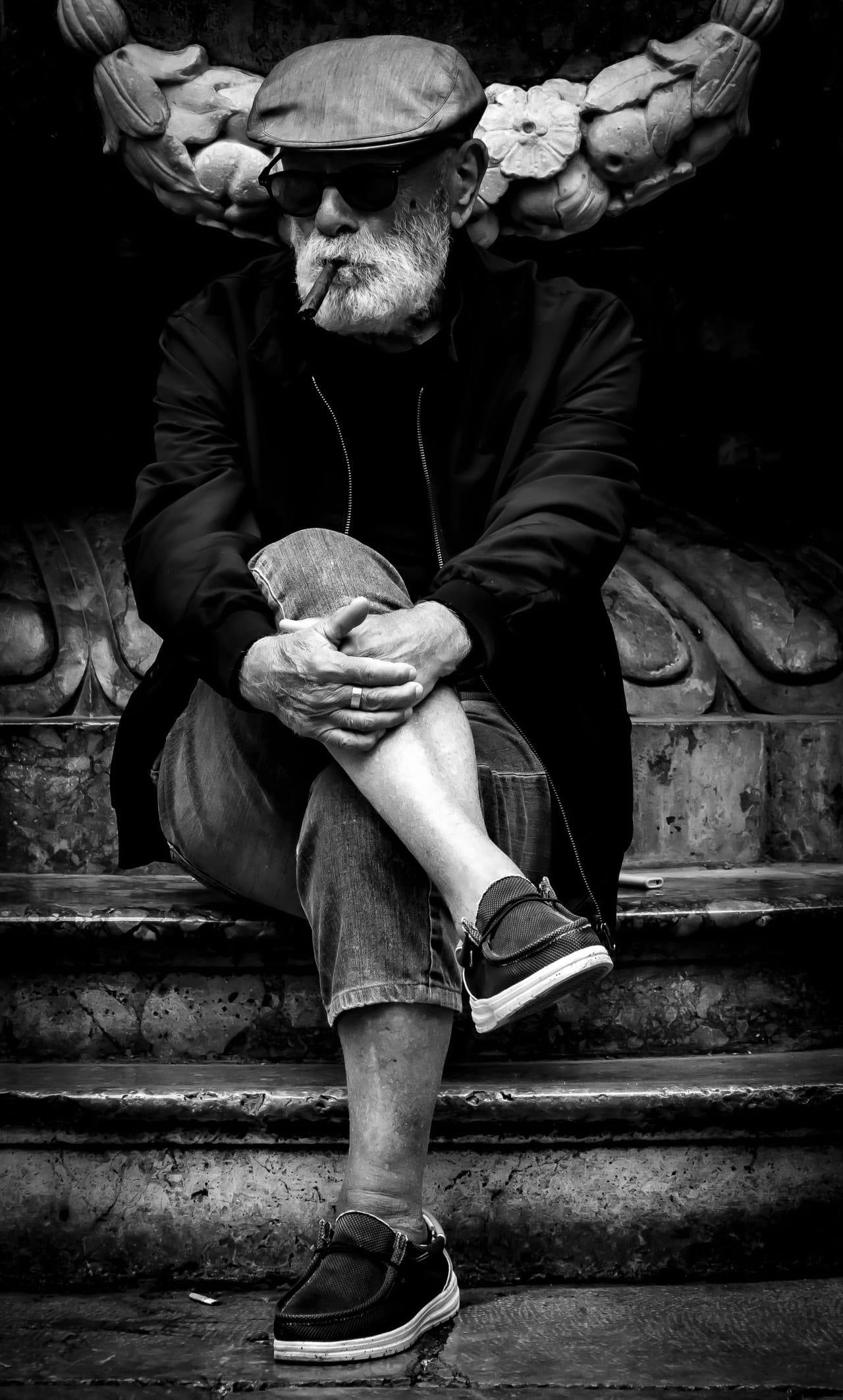 Black-and-white photo of an older bearded man wearing a flat cap and dark sunglasses, sitting on worn stone steps with one leg crossed, hands clasped over his knee, and a cigar in his mouth; carved floral stonework fills the background.