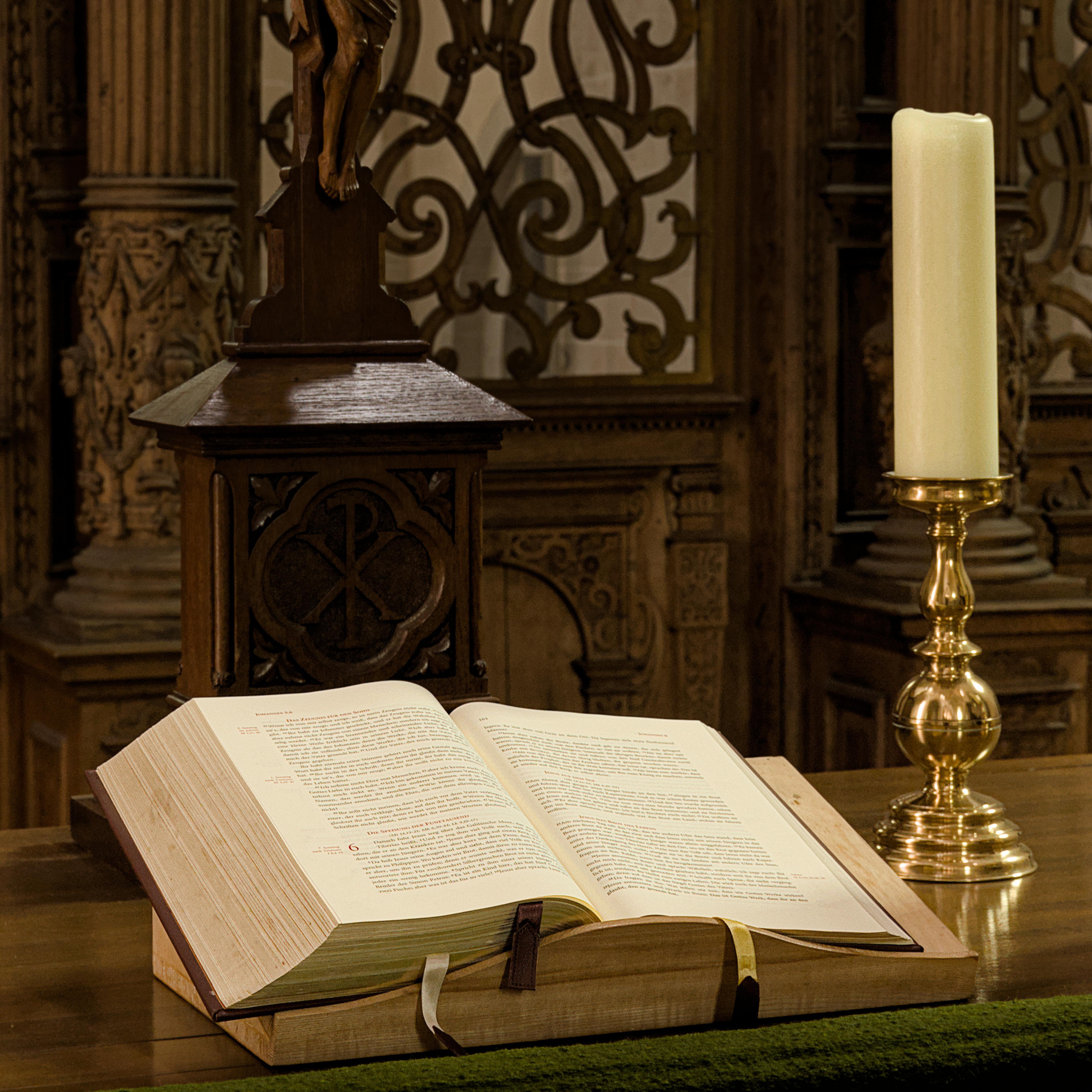 Open book on a wooden lectern with intricate carvings, placed on a wooden surface. A tall, unlit white candle stands in a gold candlestick nearby. Background features ornate wood detailing and scrollwork. 
### 
Offenes Buch über ein hölzernes Rednerpult mit aufwendigen Schnitzereien, die auf einer Holzoberfläche platziert sind. Eine große, unbeleuchtete weiße Kerze steht in einem goldenen Kerzenständer in der Nähe. Hintergrund zeigt kunstvolle Holzdetails und Scrollwork. 