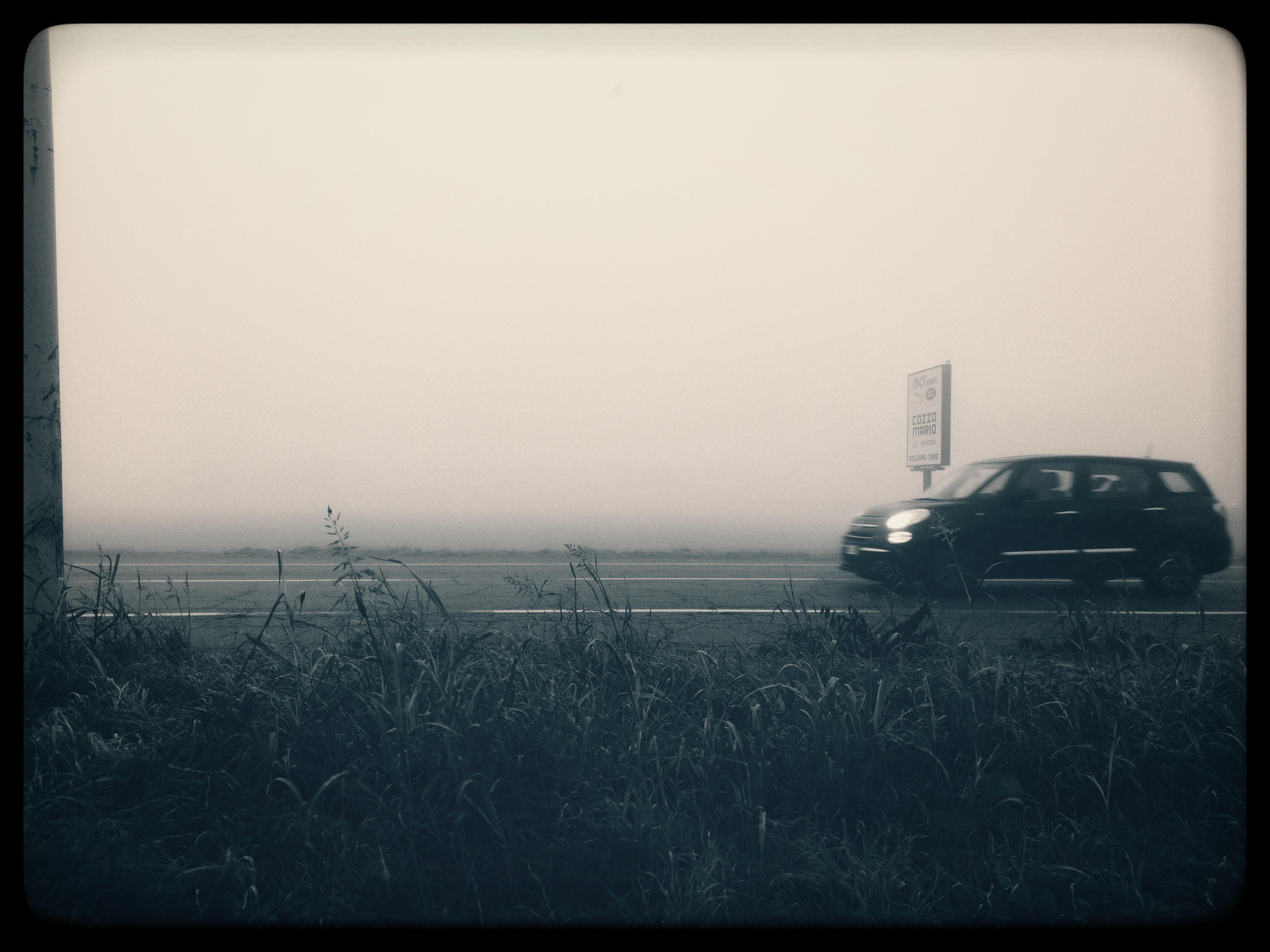 The image shows a black and white scene with a car driving on a road. There is a grassy area in the foreground, and a sign is visible near the road. The background appears foggy, creating a misty atmosphere.