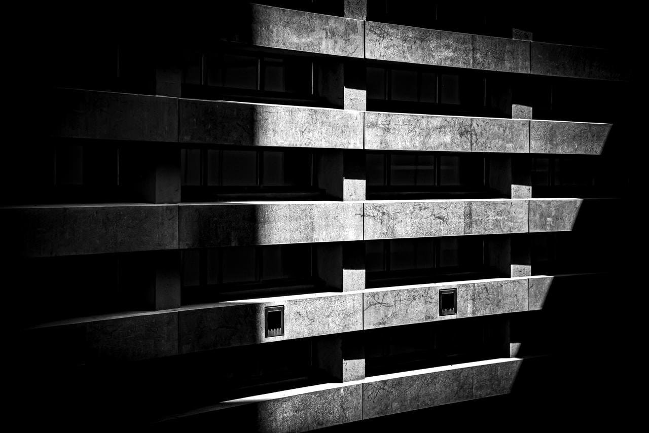 Black and white image of a concrete building with horizontal rows of windows. Diagonal shadows create a dramatic pattern across the facade, highlighting the building's geometric structure. The image has high contrast with deep shadows and bright highlights on the concrete surface.