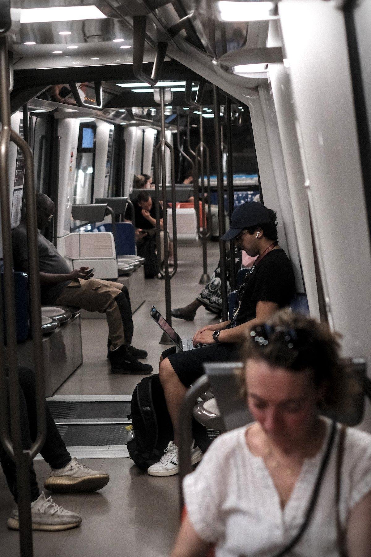 Photo verticale. Dans une rame de métro, un homme en t-shirt, short et casquette noirs, pianote sur son ordinateur portable posé sur ses genoux