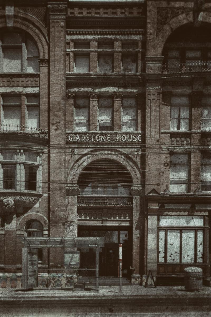 Front facade of a multi-story brick building with arched entryway, rows of tall windows, and decorative stonework; a sign above the arch reads “GADSTON HOUSE,” with sidewalk barriers and utility wires in the foreground.