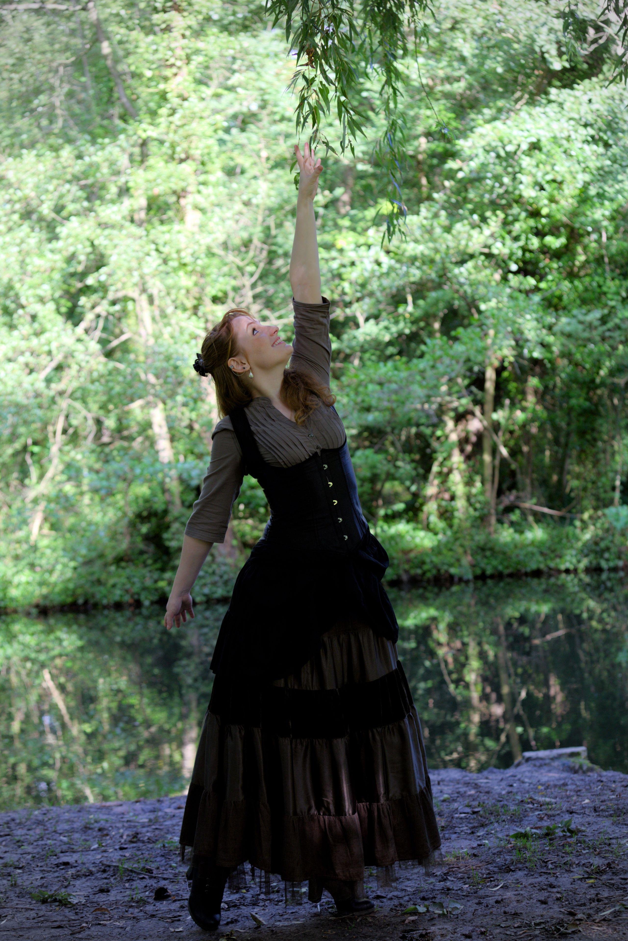 Photo portrait, montrant une jeune femme rousse au cheveux long élégamment coiffé, portant un chemisier marron et par dessus, un corset noir, une longue jupe marron foncé. Elle se tiens sur la pointe des pieds et essai de toucher une branche d’arbre loin au dessus d’elle. Le fond est composé d’un plan d’eau calme et d’arbres