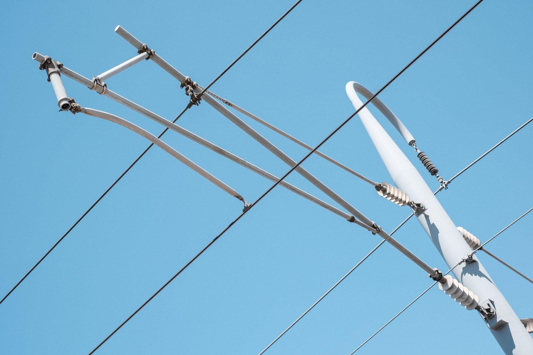 Photo horizontale. Sur fond de ciel bleu impeccable, des lignes électrique de train parallèles passant par une espèce de ceintre sur un poteau gris clair presque blanc