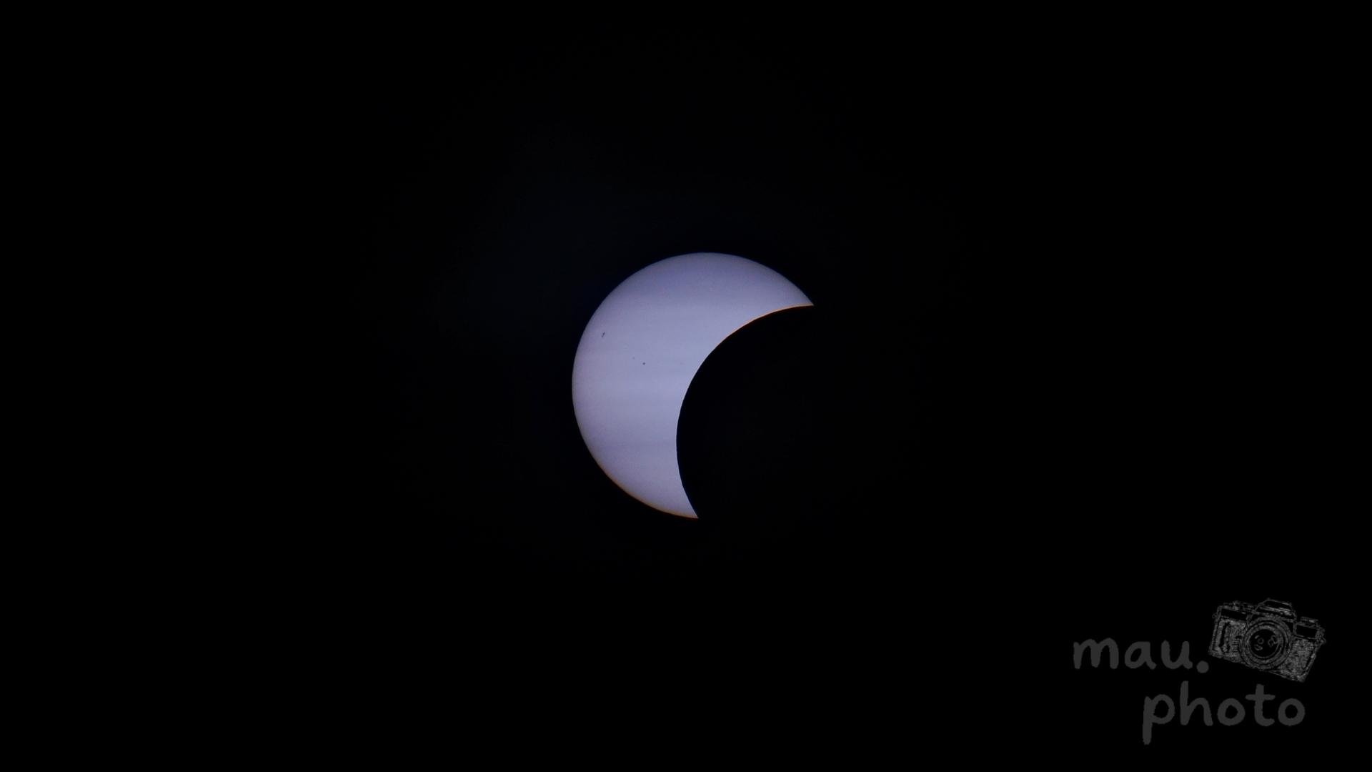 Partial solar eclipse with the sun appearing as a bright white crescent against a black sky, centered in the frame, with a “mau.photo” watermark in the bottom right.