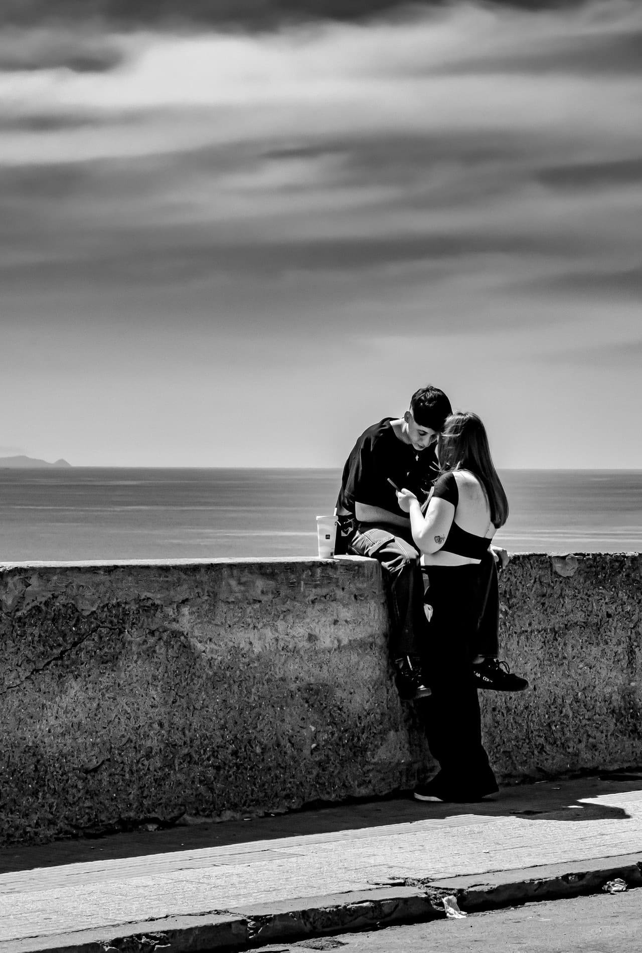 Black-and-white photo of two people leaning against a low concrete seawall, heads close together as they look down at something in their hands, with the ocean and a cloudy sky in the background and a paved walkway in the foreground.