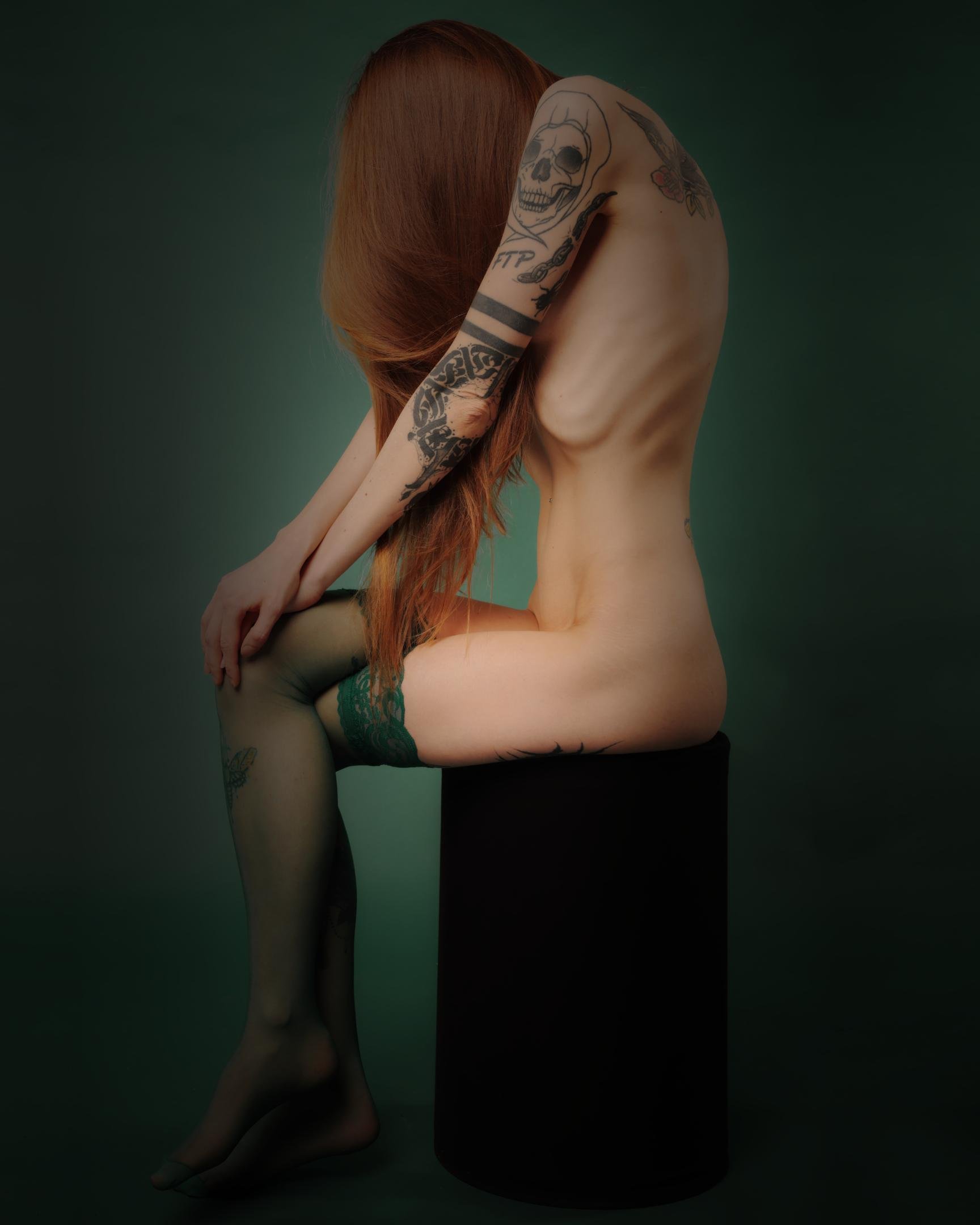 Naked, dressed only in green stockings girl. We see a left side of her, sitting on black waltz chair. She is thin and has tattoo on her left shoulder.