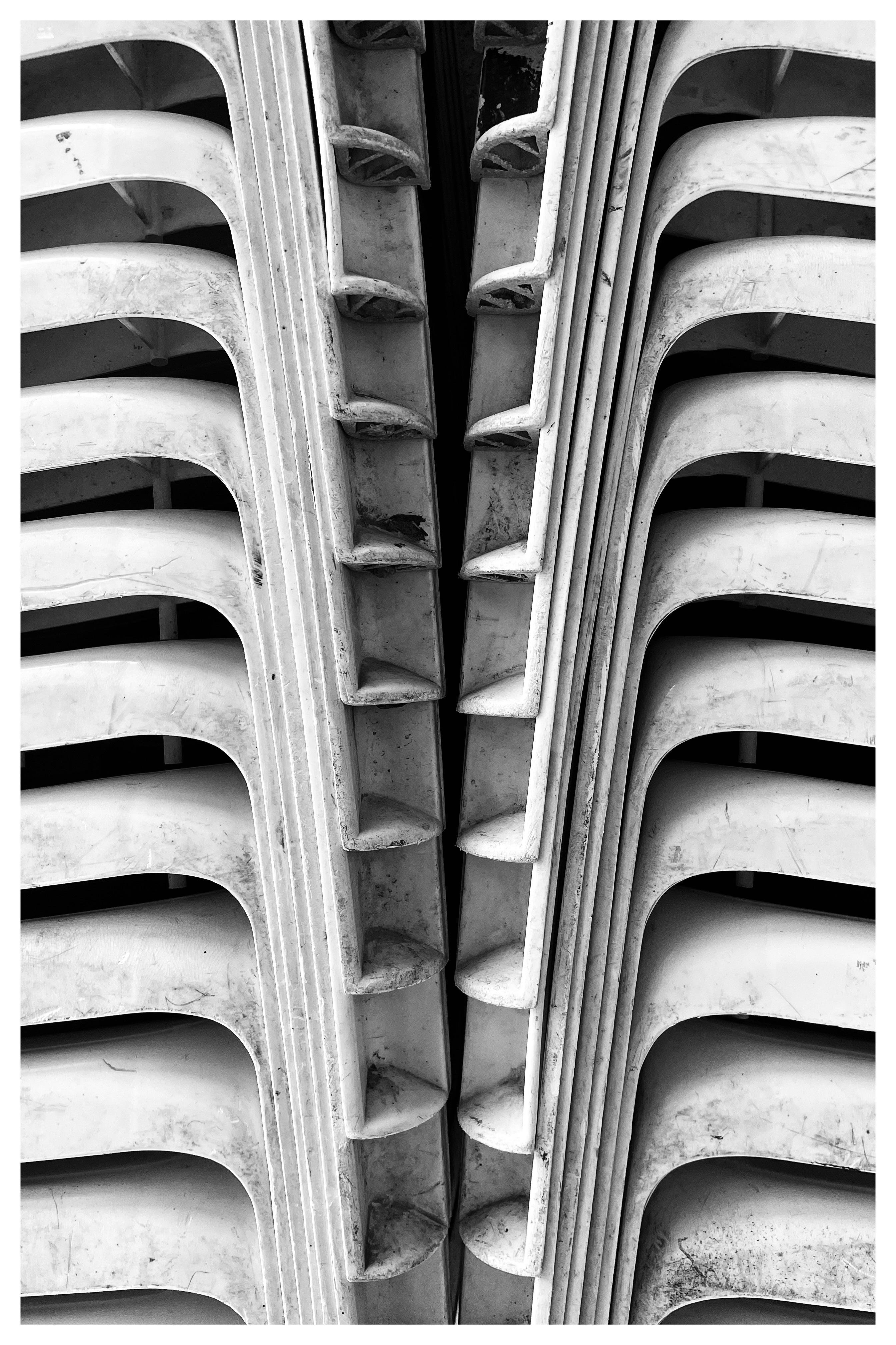 Close-up, black-and-white view of two stacks of nested white plastic chairs facing each other, with curved seat edges and vertical slats forming a narrow dark gap in the center.