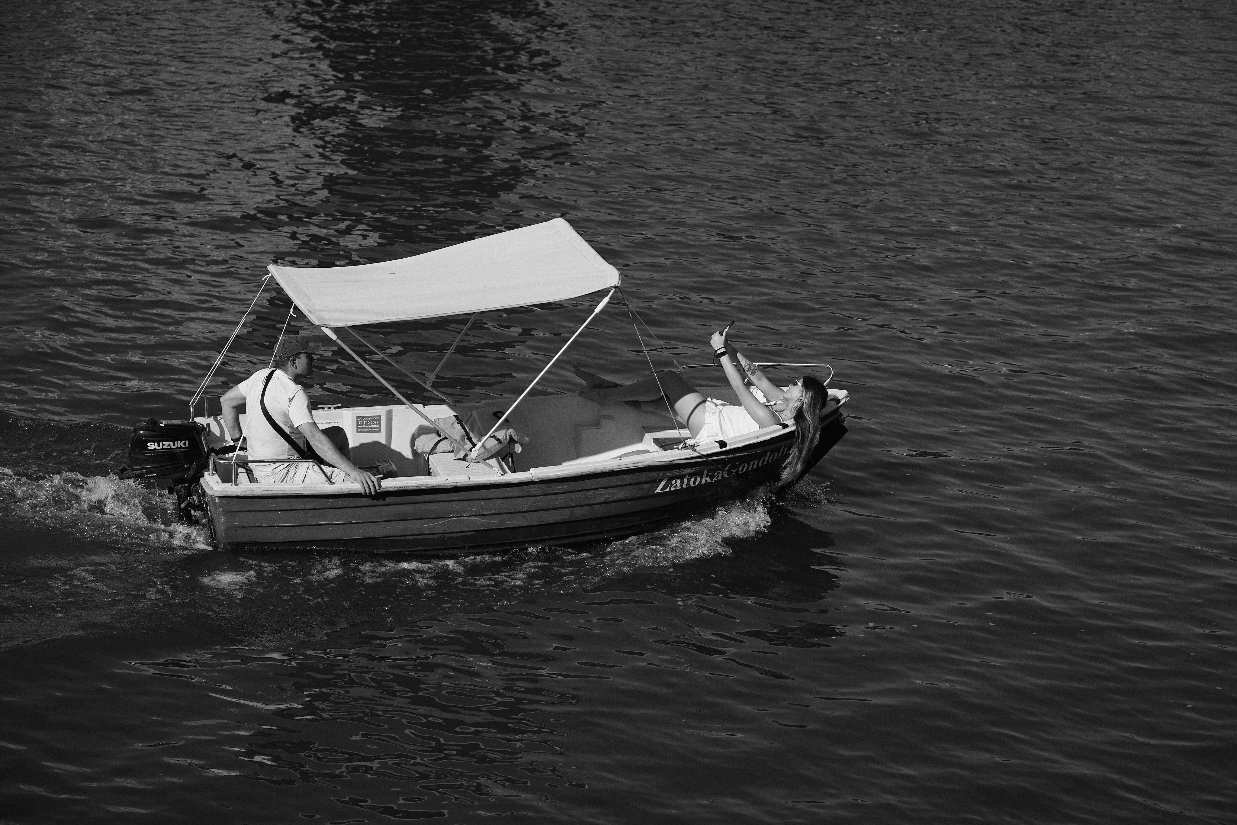 Motorboat with a canopy on open water, carrying two people. One person sits at the back operating the boat, while the other reclines at the front, holding up a smartphone. Visible text on the boat's side. Black and white filter applied.