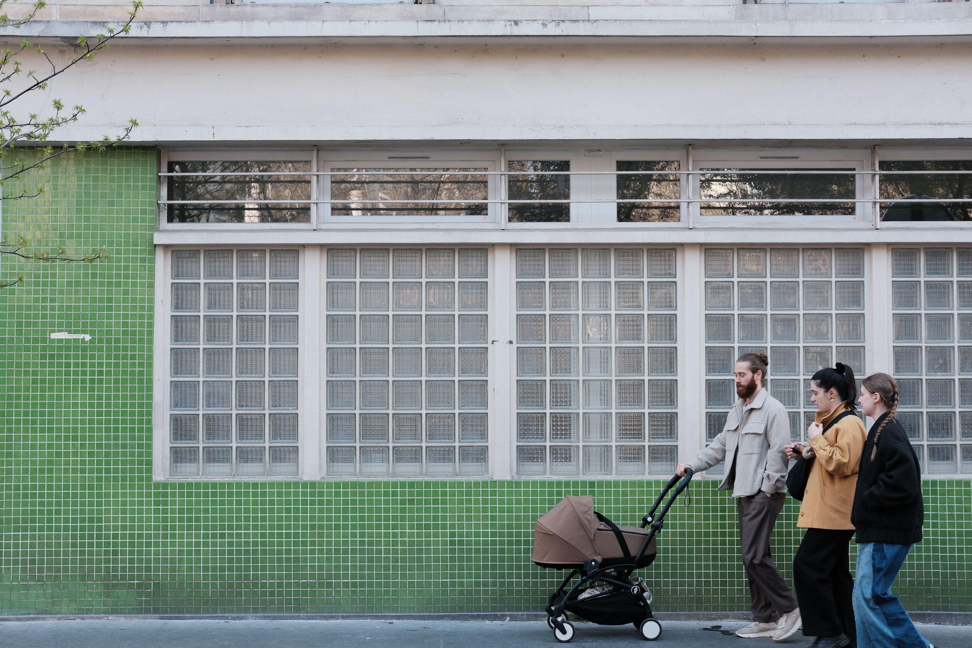 Photo horizontale. Façade d’un bâtiment avec des fenêtres faites de petits carreaux carrés et brouillés et le mur couvert de petite céramiques vertes carrées. 3 personnes, un homme et 2 femmes, passent devant depuis la droite, l’homme poussant une poussette
