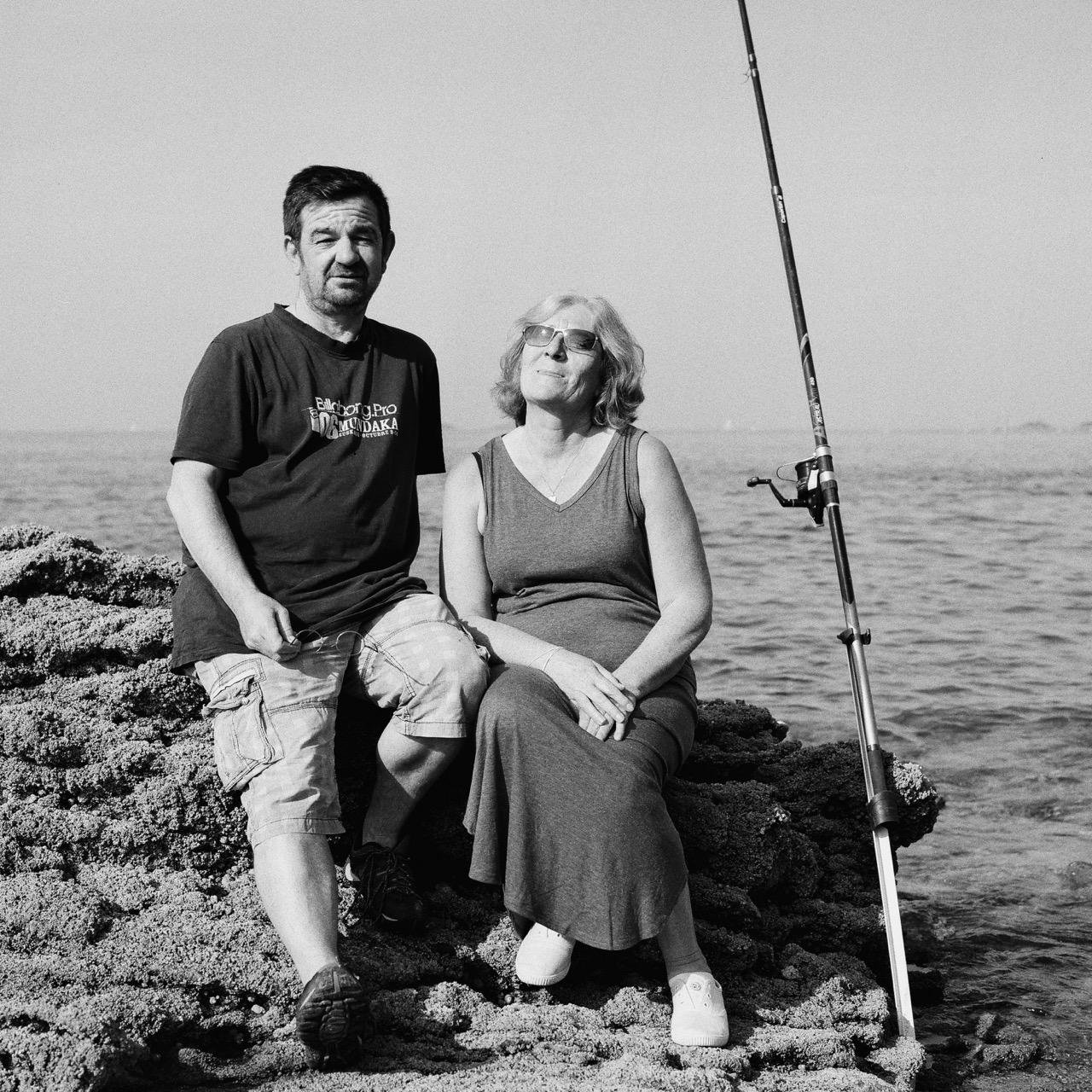 The image shows a black and white photograph of two people sitting on rocks by the sea. A man is on the left wearing a t-shirt and shorts, and a woman is on the right wearing a dress. Next to them is a fishing rod. It's a serene scene by the water.