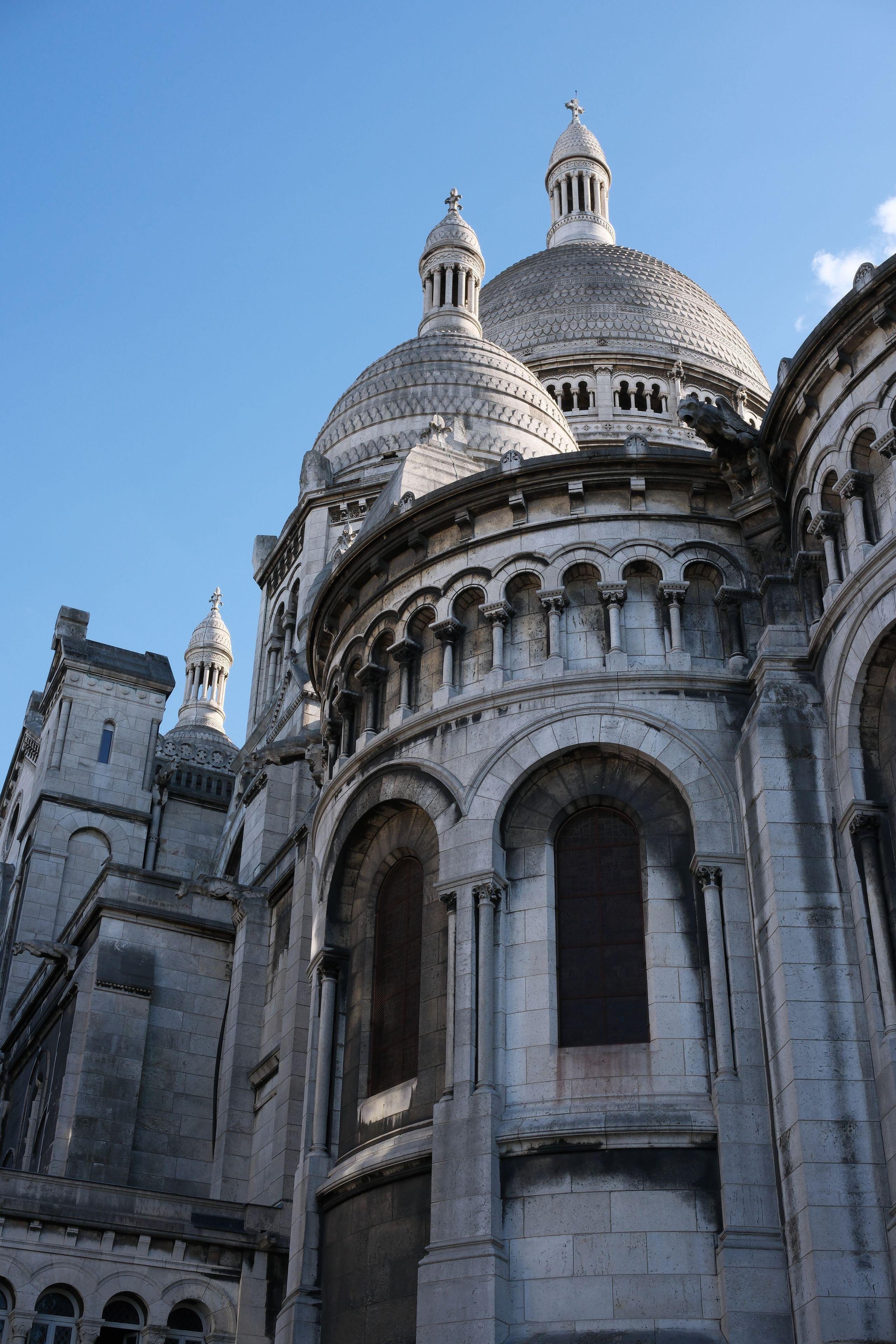 Photo verticale. Vue en contre-plongée sur le Sacré-Cœur sous un ciel bleu d’été sans nuage. La façade est dans l’ombre faisant ressortir la crasse accumulée sous les reliefs de la bâtisse