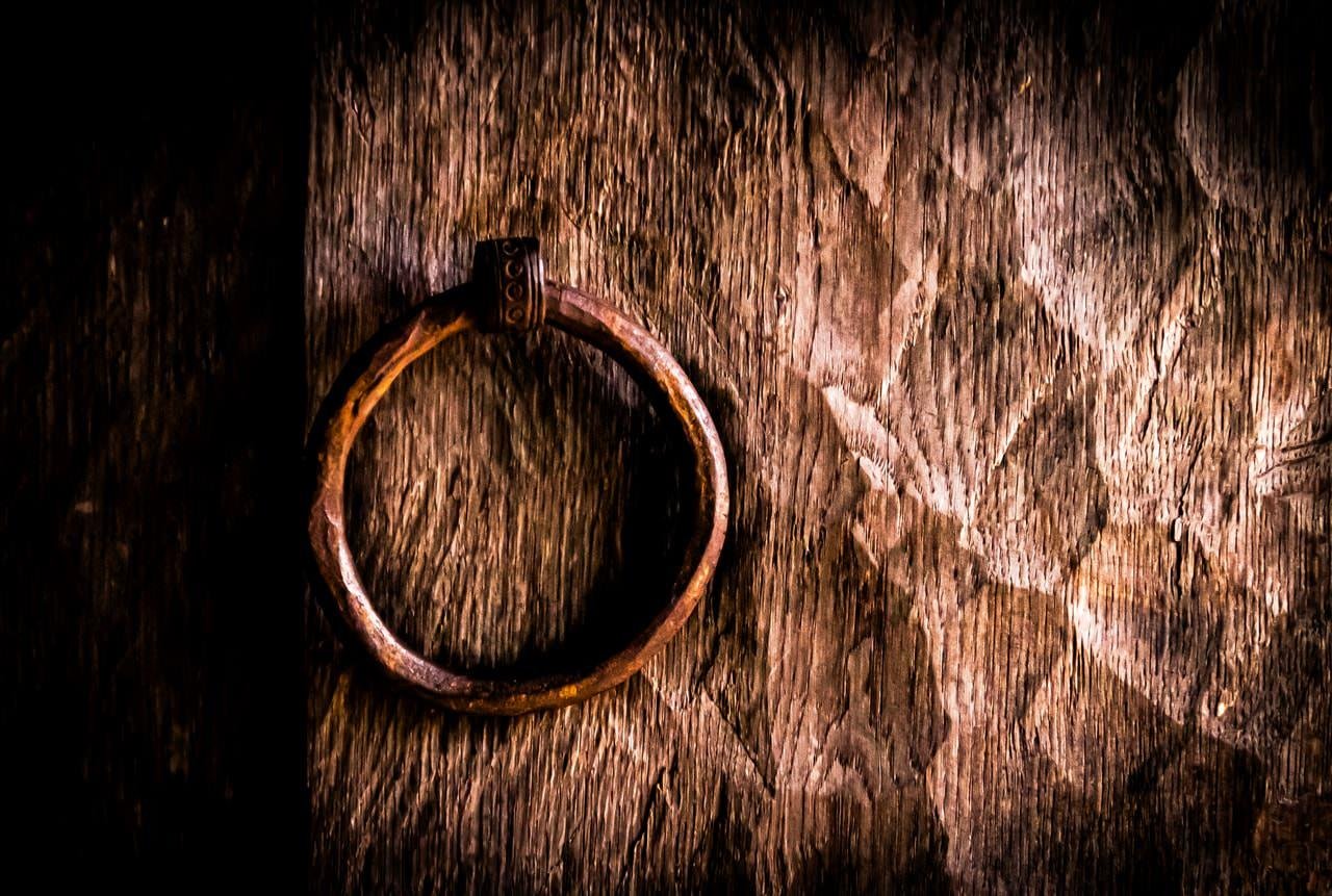 A weathered, circular metal ring attached to a textured, rough wooden door. The wood displays dark, uneven patches and visible grain patterns, with the ring placed to the left. The lighting creates a dramatic contrast, emphasizing the wood's rugged surface.