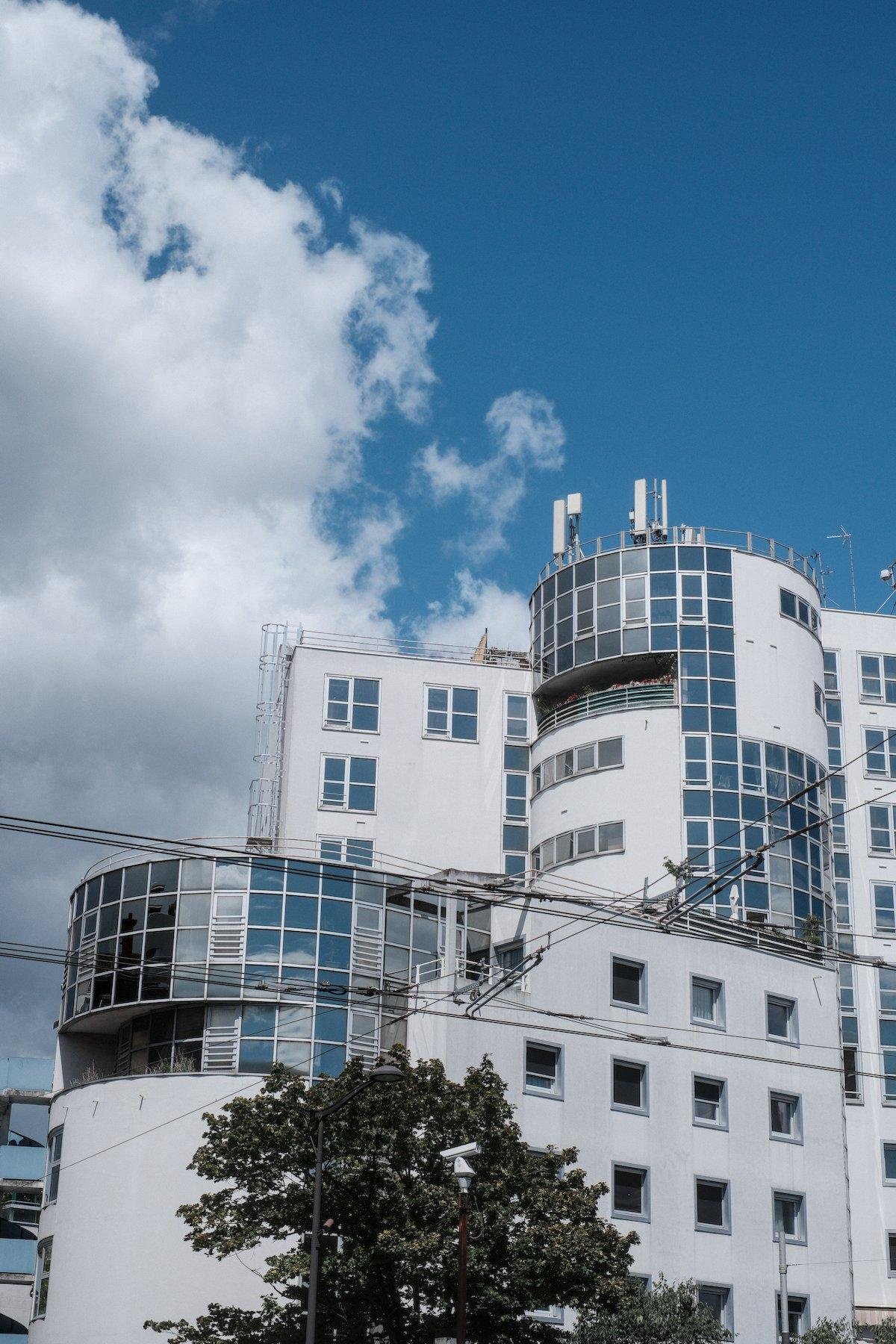 Photo verticale. Un immeuble blanc majoritairement rectangulaire, mais avec 2 parties cylindrique avec des baies vitrées, sous un ciel bleu avec un gros nuage à gauche de l’image