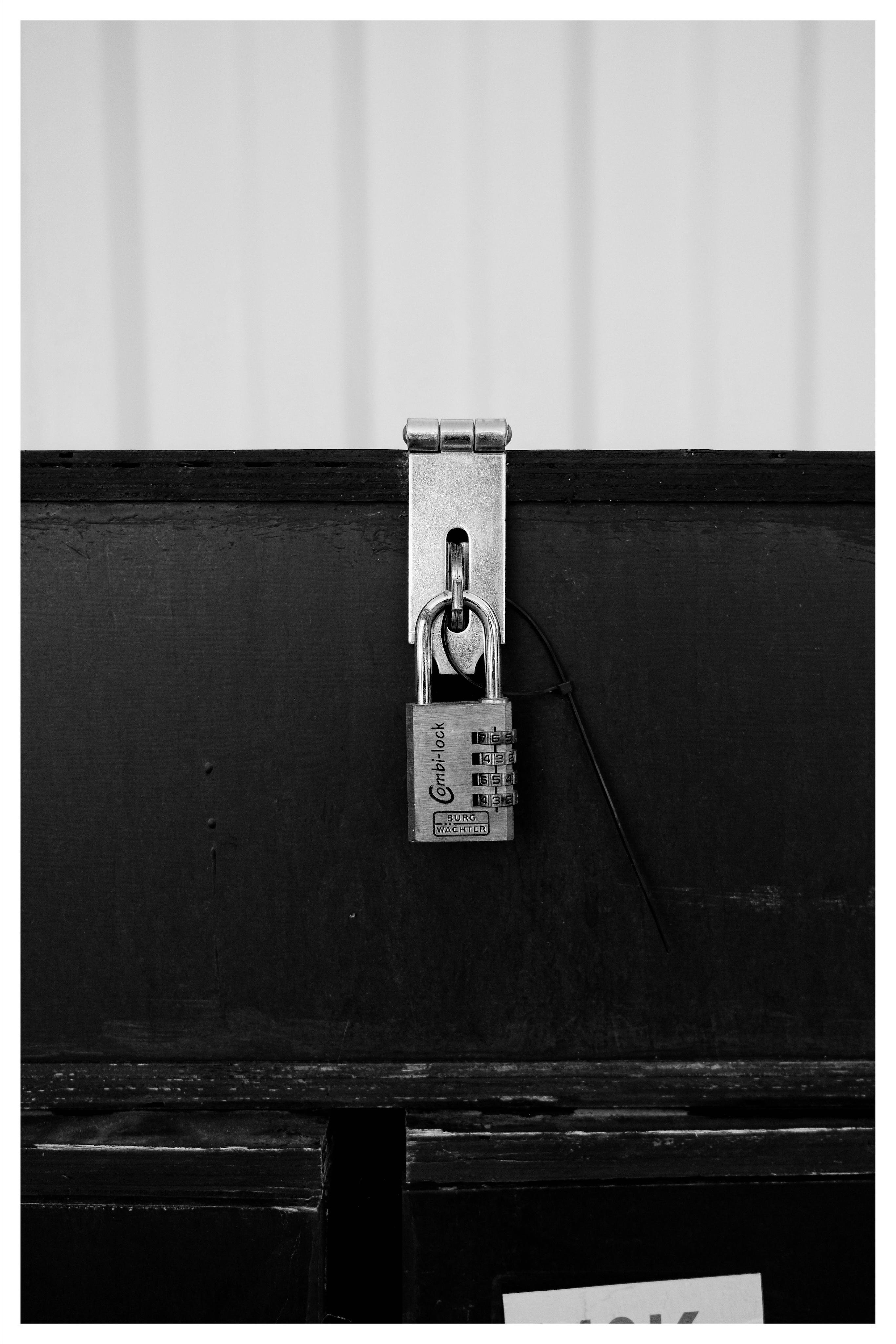 Silver combination padlock hanging from a metal latch on a dark wooden box, centered in a black-and-white close-up.