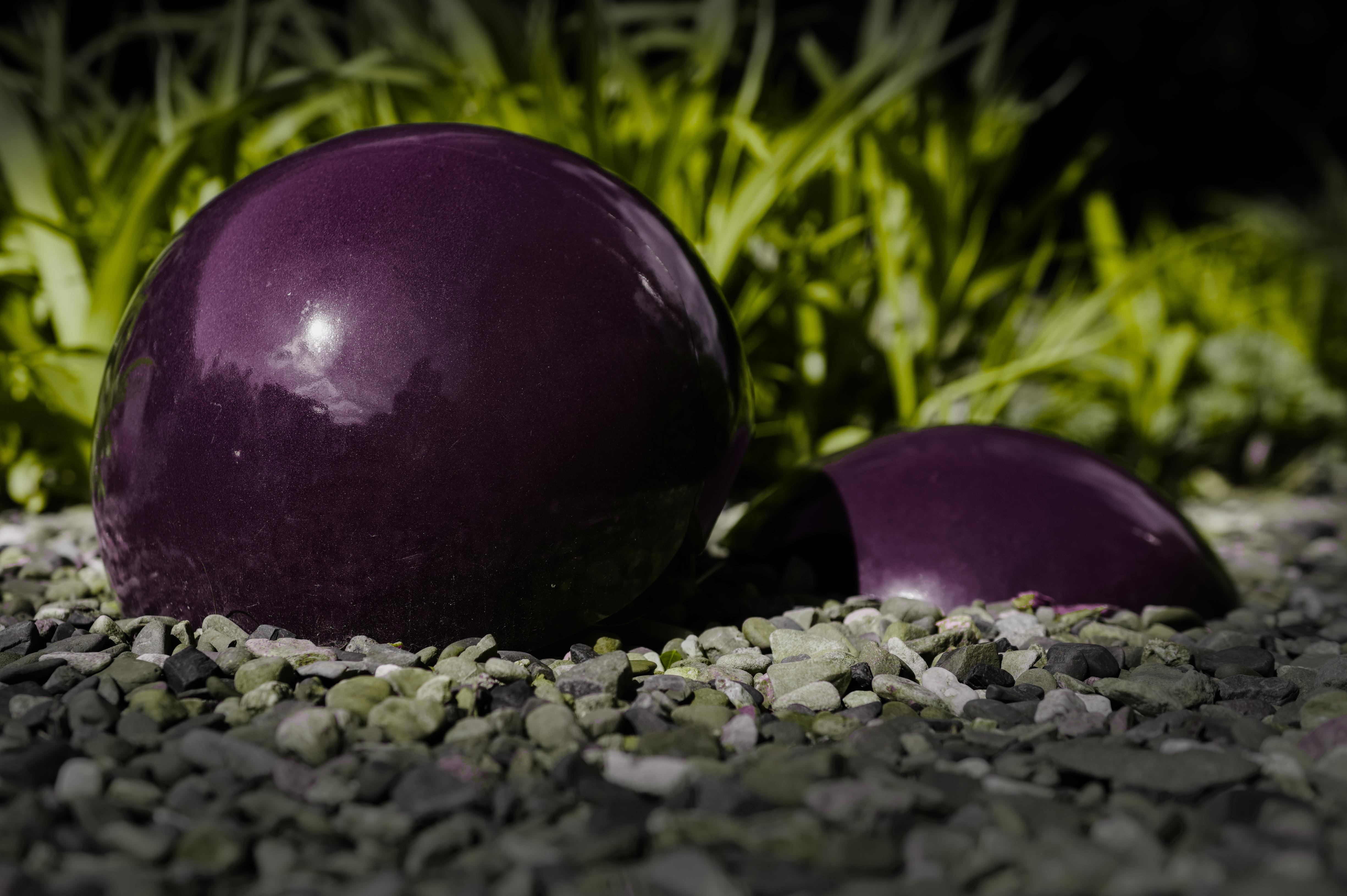 Two glossy dark-purple spheres resting on a bed of small gray gravel, with green plants blurred in the background. 
### 
Zwei glänzende, dunkelviolette Kugeln ruhen auf einem Bett aus kleinem grauen Kies, im Hintergrund sind grüne Pflanzen verschwommen zu erkennen.
