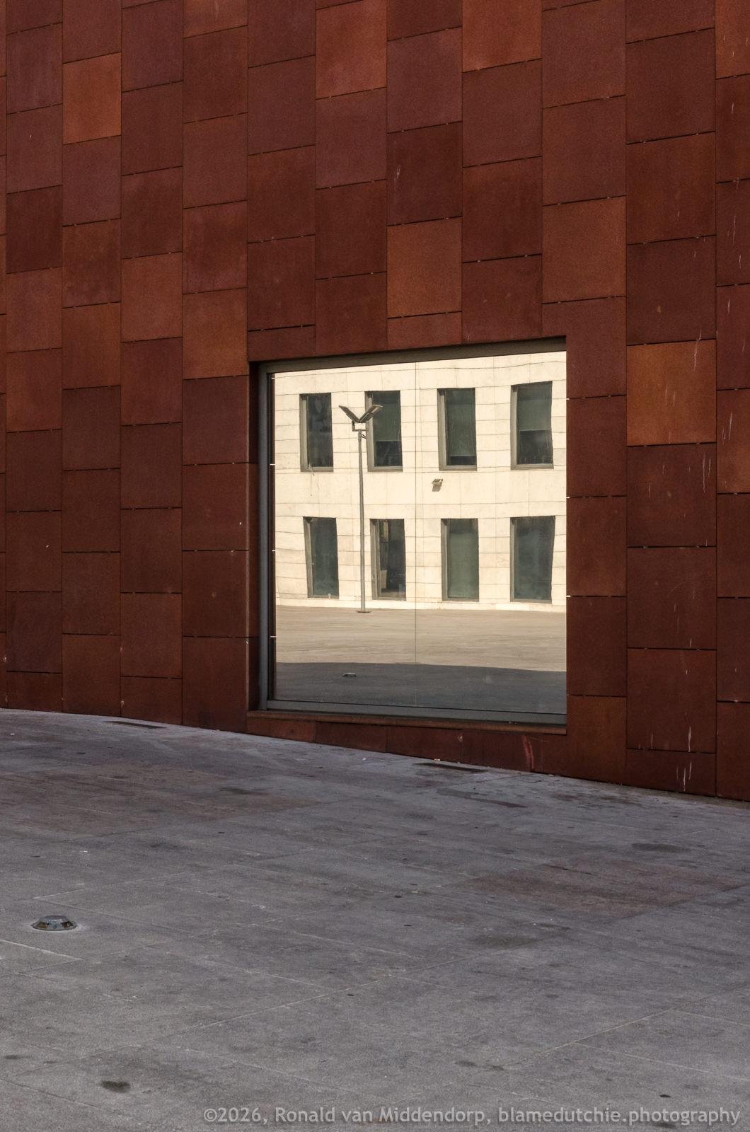 Reddish-brown wall with a large rectangular reflective window near the bottom center, mirroring a pale building with tall windows and a courtyard lamp post; gray concrete ground in the foreground.