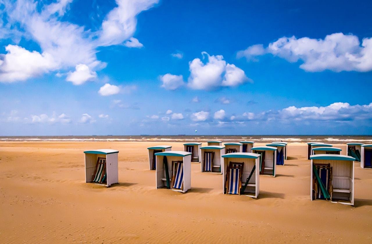 The image shows a beach with several empty beach huts or cabanas placed on the sand. They are facing towards the ocean under a blue sky with scattered clouds.